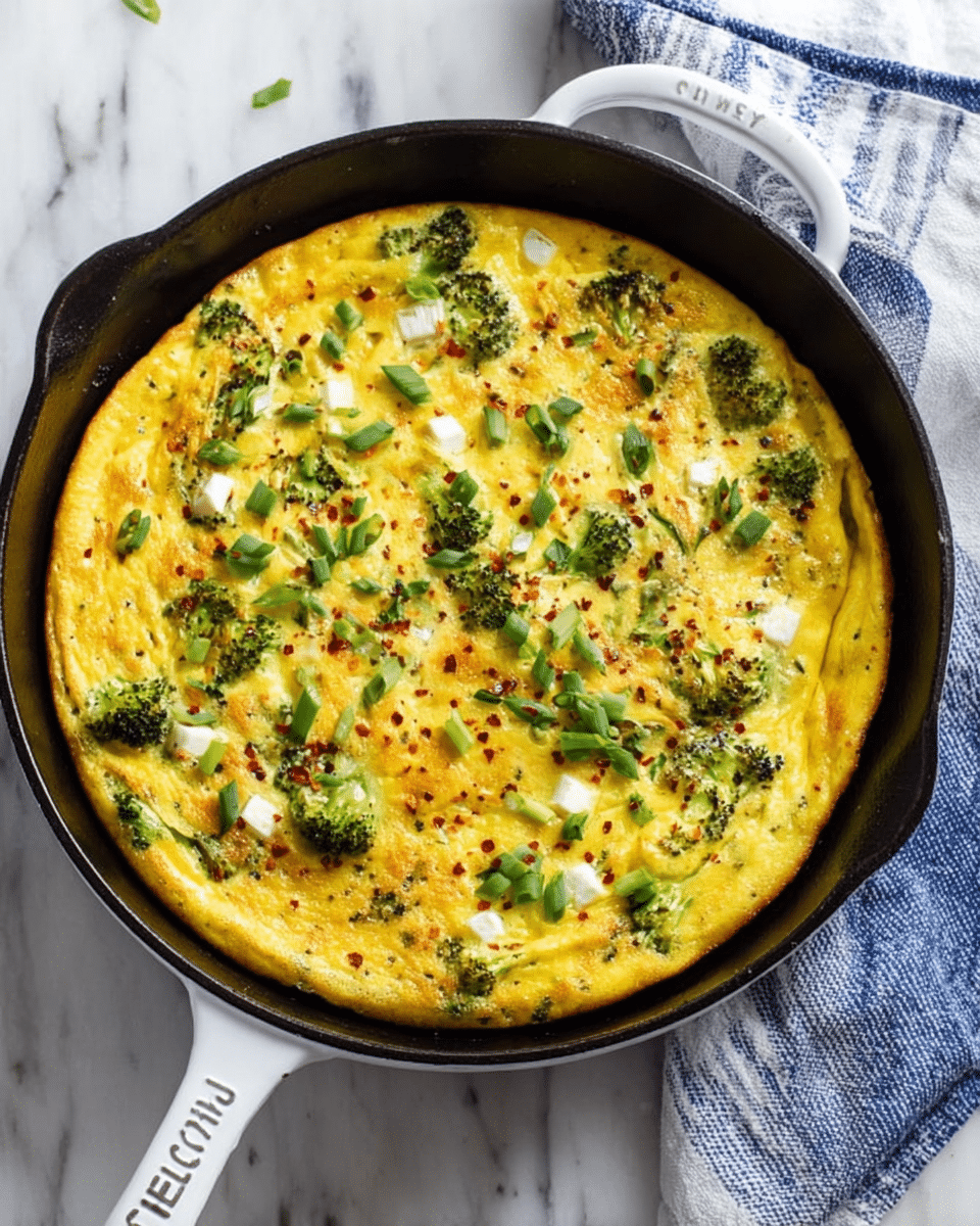 The image shows a golden-yellow frittata cooked in a black cast iron skillet, placed on a white marbled surface with a blue and white striped cloth near the handle. The frittata has a smooth, slightly raised texture with scattered small pieces of white cheese melting on top. Sprigs of green broccoli and chopped green onions are spread evenly across the surface, along with a light sprinkling of red chili flakes adding tiny red spots. The skillet has a white handle with the brand name visible, and the dish looks fresh and vibrant. Photo taken with an iphone --ar 4:5 --v 7