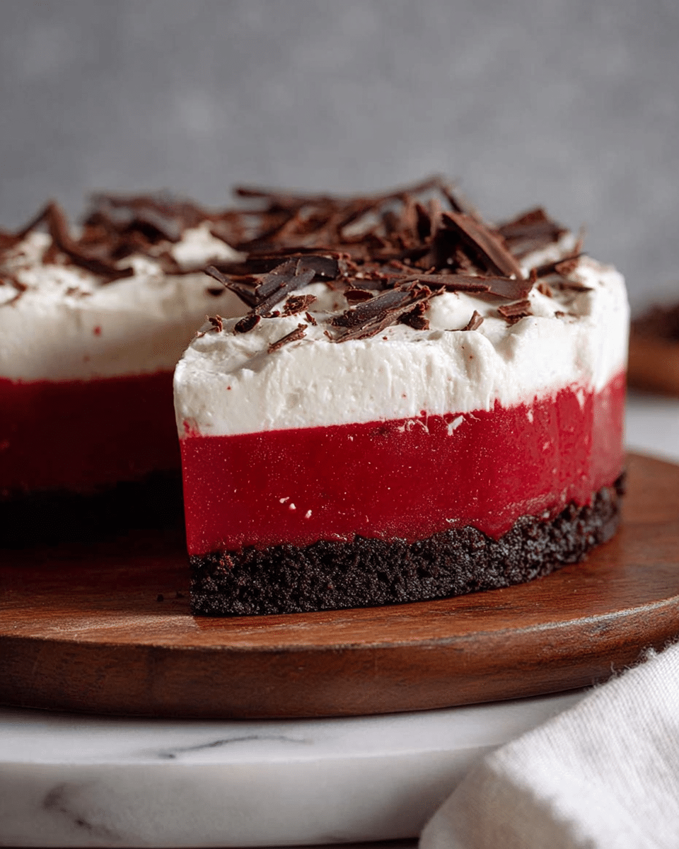 The image shows a three-layer dessert with a dark, crumbly base layer that looks like chocolate cookie crust, topped by a smooth, bright red jelly layer in the middle, and finished with a thick, fluffy white cream layer on top. The cream layer is sprinkled with thin dark chocolate shavings. The dessert is placed on a wooden stand against a soft gray background, and a white marbled surface with a white cloth is partially visible in the lower area. photo taken with an iphone --ar 4:5 --v 7
