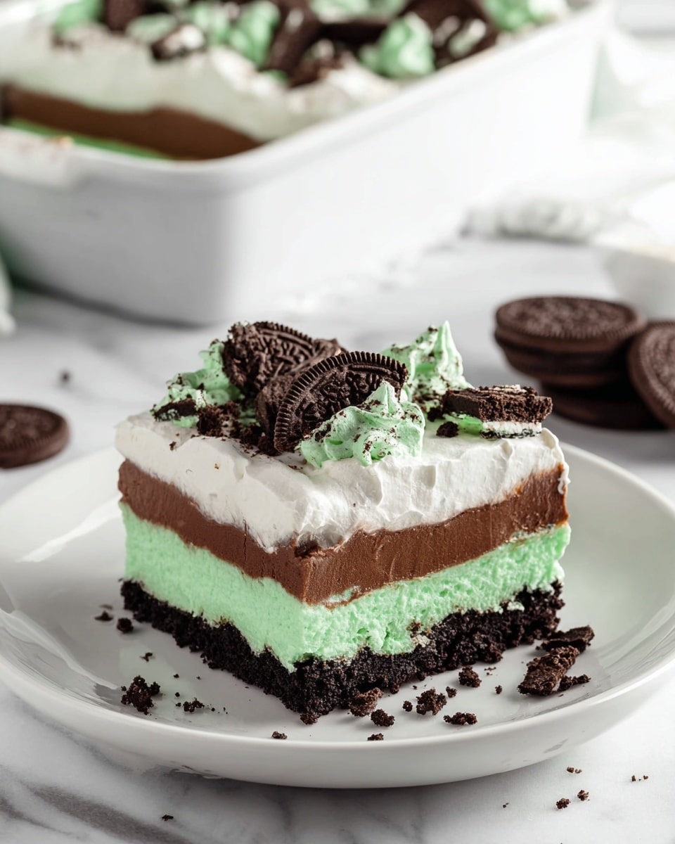 A square piece of layered dessert sits on a white plate placed on a white marbled surface. The dessert has four visible layers: the bottom layer is a dark, crumbly chocolate crust, the second layer is a smooth, bright green mint cream, the third layer is a thick, rich chocolate pudding, and the top layer is a fluffy white whipped cream. On top of the whipped cream are scattered broken pieces of chocolate cookies with green cream filling, adding texture and rich dark and green colors. Cookie crumbs are scattered around the plate. In the background, a white rectangular baking dish filled with the same dessert and extra whole chocolate mint cookies can be seen. photo taken with an iphone --ar 4:5 --v 7