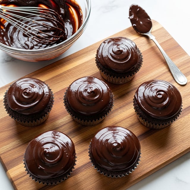 The image shows six chocolate cupcakes arranged on a wooden cutting board with a warm brown tone. Each cupcake has one smooth, thick layer of glossy dark chocolate frosting on top that reflects light softly, covering the entire top surface with slight swirls and peaks. The cupcakes sit in silver foil liners, and the texture of the cake underneath appears moist and dark. Nearby, there is a clear glass bowl filled with more glossy chocolate frosting, with a metal whisk resting inside it. A silver spoon with chocolate on it rests on the upper right corner of the board. The background is a white marbled texture. Photo taken with an iphone --ar 4:5 --v 7