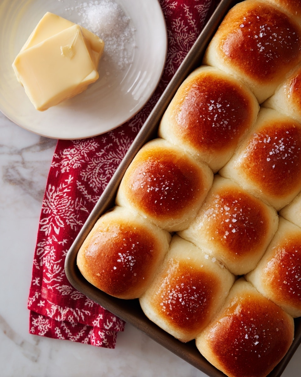 This image shows a rectangular metal baking pan filled with twelve soft, golden brown dinner rolls arranged in three rows and four columns. Each roll has a slightly shiny, crisp crust on top with a warm, light golden color, and soft, fluffy texture visible on the sides. The rolls are touching each other closely, making the edges slightly pressed together. One roll in the middle of the top row is tilted outward, revealing its soft, pale interior. The pan sits on a white marbled surface with a red and white zigzag patterned cloth partially visible in the top left corner. photo taken with an iphone --ar 4:5 --v 7
