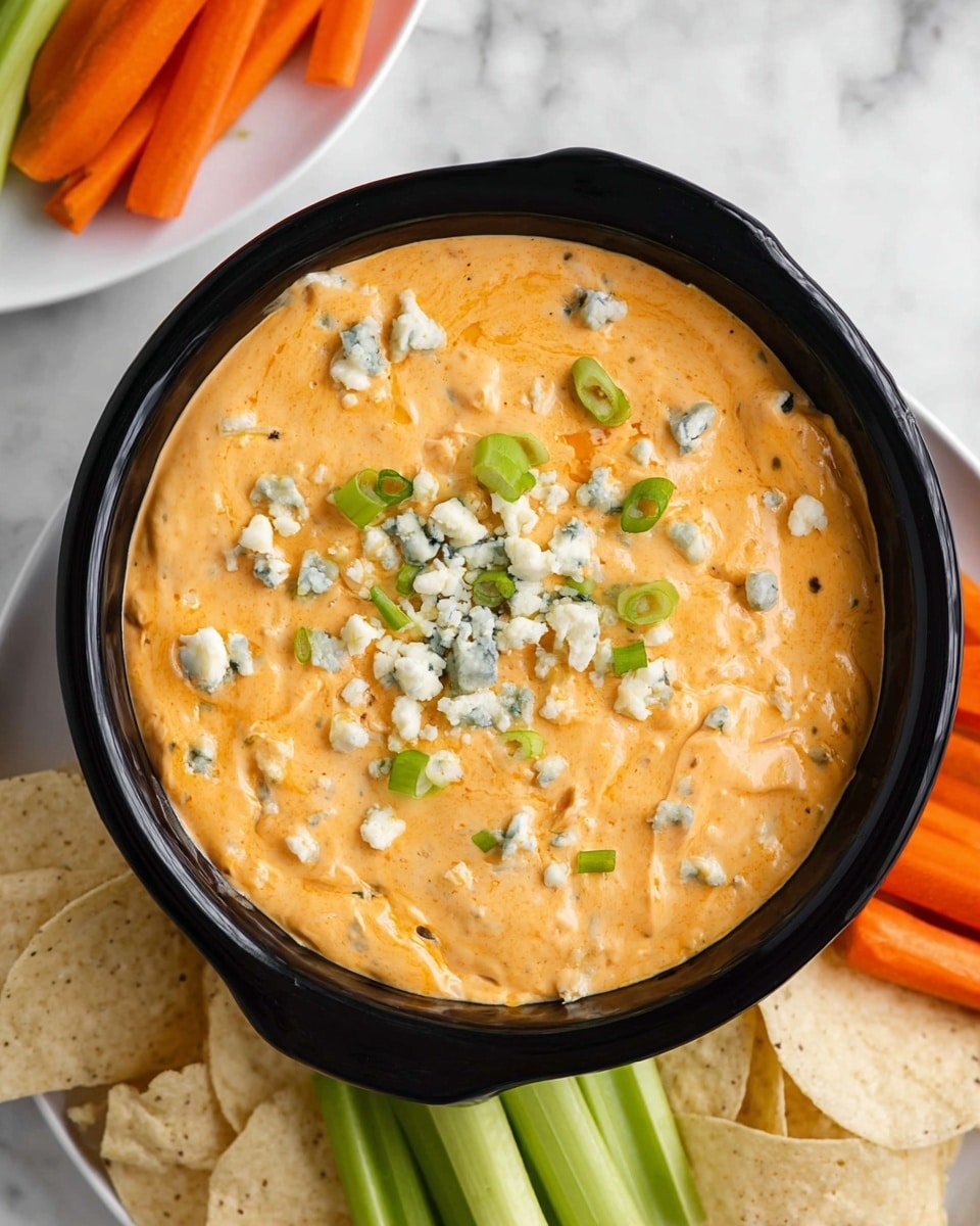 A creamy orange dip fills a black bowl, topped with small chunks of white and blue-speckled cheese and scattered bright green sliced scallions. The dip has a smooth, slightly chunky texture. Around the bowl, there are creamy beige tortilla chips and a white plate holding bright green celery sticks and fresh orange carrot sticks. The setting is on a white marbled surface. photo taken with an iphone --ar 4:5 --v 7