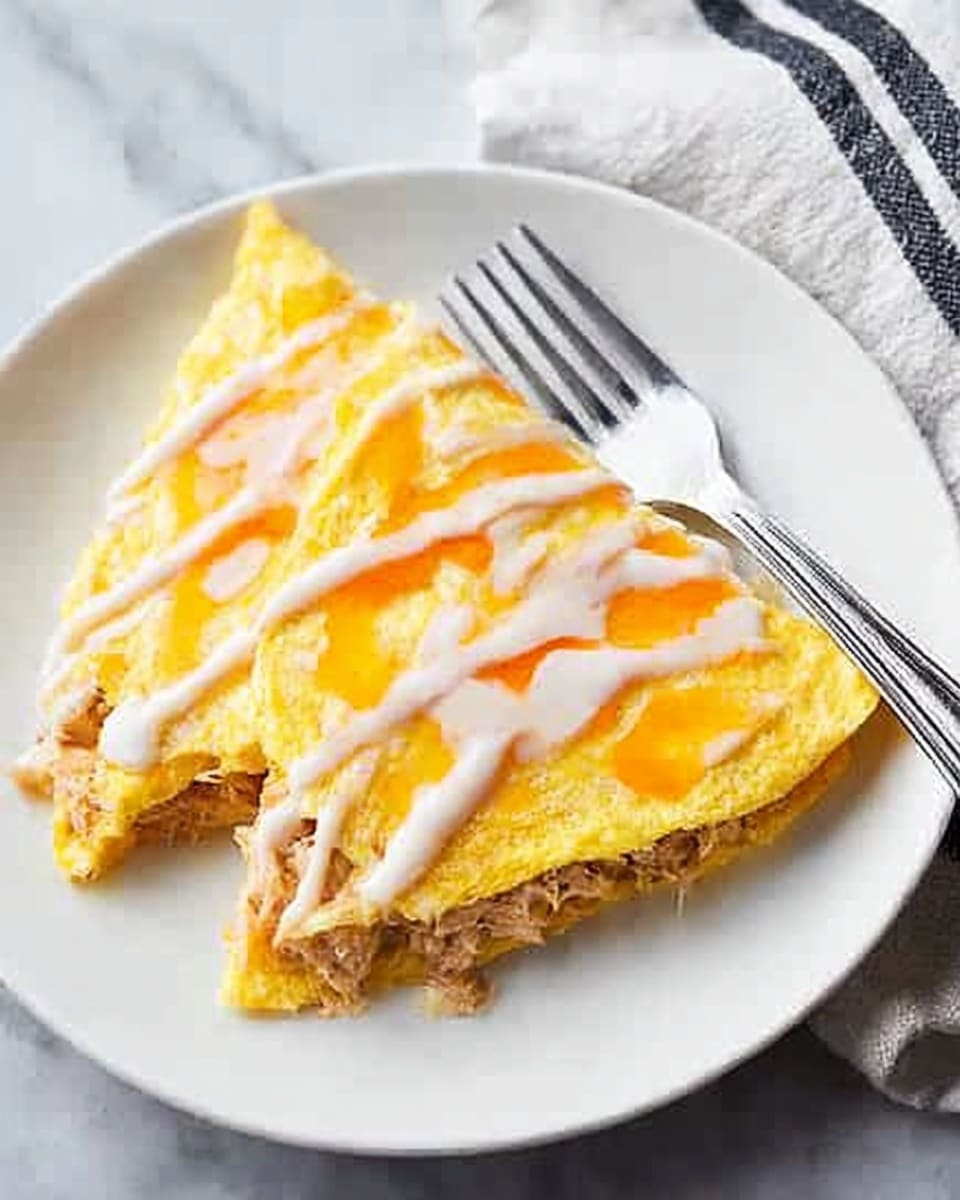 A close-up image of a white plate holding two folded, yellow omelette slices with a soft, slightly browned texture, filled with light brown shredded meat visible inside. The top of the omelettes is covered with melted cheese in a pale yellow shade. White sauce is drizzled in thin lines over the omelettes, along with an orange sauce creating a contrast on top. A silver fork rests on the right side of the plate, placed on a white marbled surface next to a white cloth with two black stripes. Photo taken with an iphone --ar 4:5 --v 7