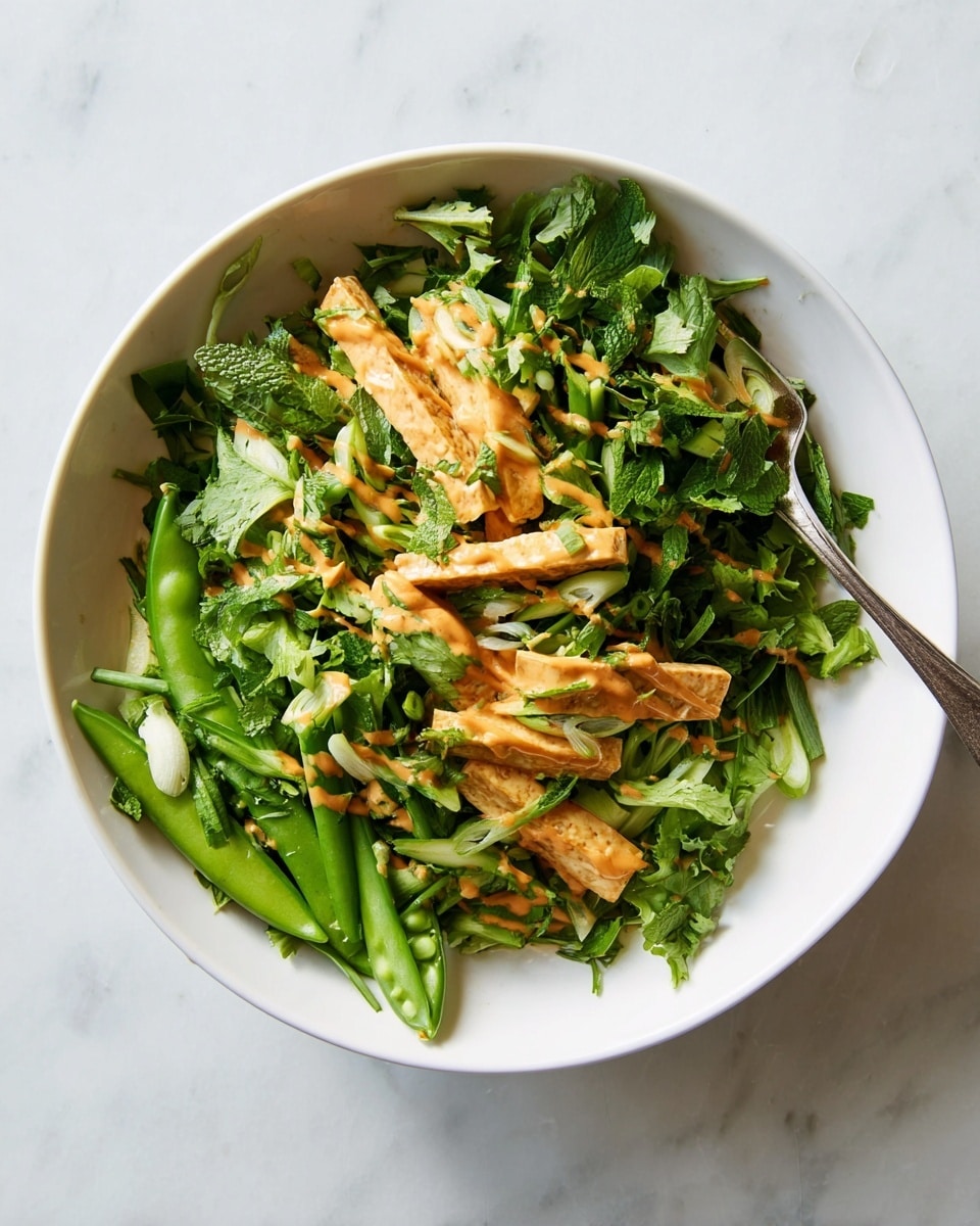 A white bowl holds a fresh, light salad with three main layers: the bottom layer is bright green leafy herbs including mint and cilantro, the middle layer is crisp, angled snap peas with a fresh green color, and the top layer consists of golden-brown strips of tofu drizzled with a smooth, orange-colored dressing. Thin slices of green onion add small pops of light green throughout. A silver fork rests diagonally on the right side of the salad, partially buried in the ingredients. The bowl sits on a white marbled surface photo taken with an iphone --ar 4:5 --v 7