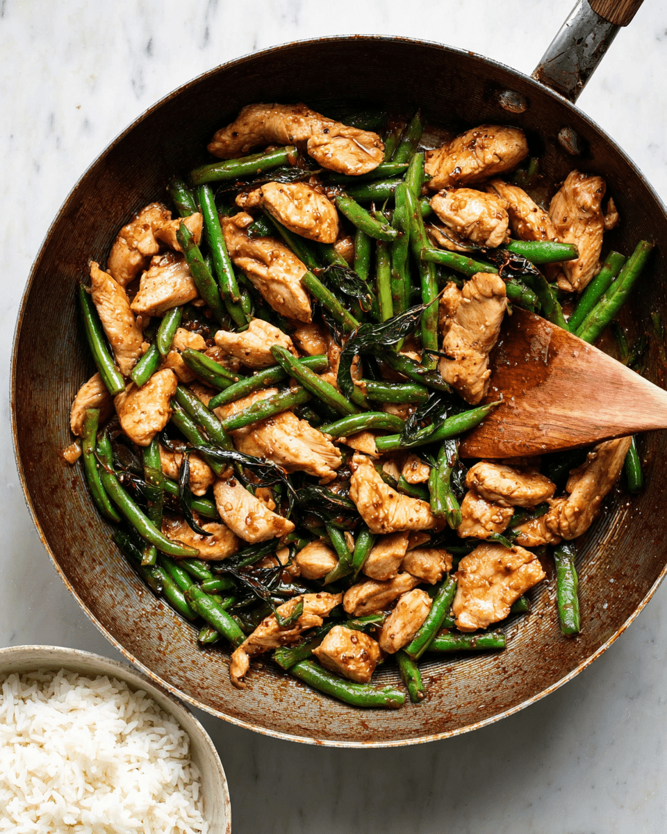 The image shows a frying pan filled with a cooked chicken and green beans stir-fry. The first layer has tender, grilled chicken pieces with a golden-brown color and a slightly shiny, saucy texture, spread evenly across the pan. Mixed among the chicken pieces are fresh green beans that are bright green with a slight wrinkled texture, showing they are cooked but still crisp. Dark green thin strips of leafy herbs are scattered throughout, adding contrast to the mix. A wooden spoon is resting inside the pan on the left side. Below the pan, there is a white bowl filled with plain white rice placed on a white marbled surface. Photo taken with an iphone --ar 4:5 --v 7