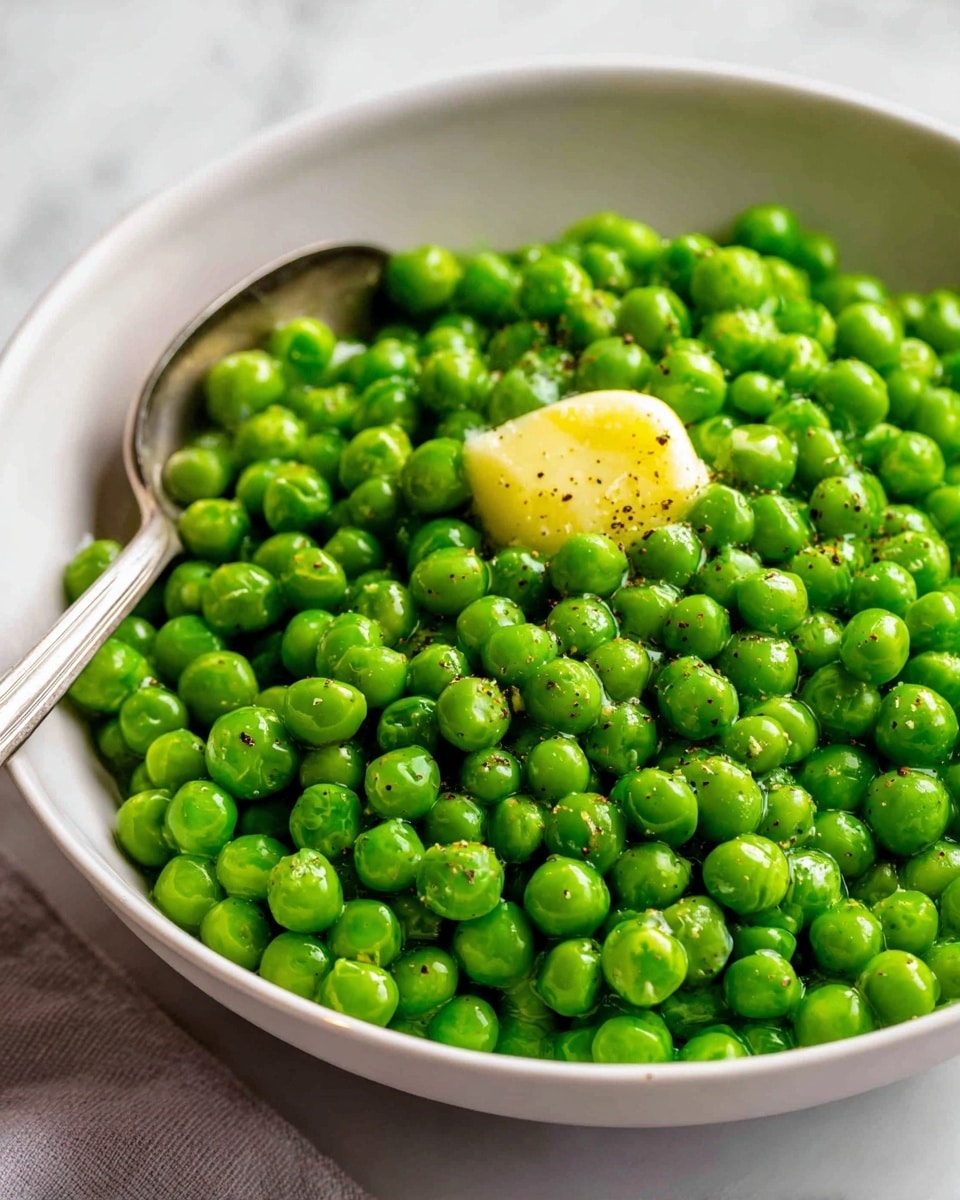 A bowl filled with bright green peas makes up the dish, with a small dollop of yellow butter melting slightly on top, sprinkled with black pepper. The peas look fresh and shiny, filling the white bowl fully, and a spoon is partially dipped into the peas on the left side of the bowl. The background is a soft white marbled texture which contrasts nicely with the green peas and the creamy yellow butter. photo taken with an iphone --ar 4:5 --v 7