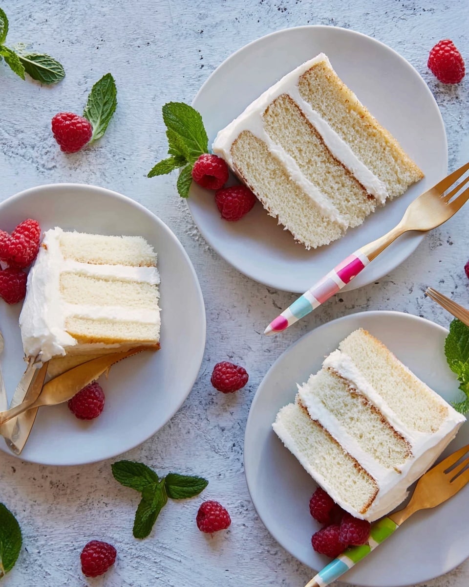 Three slices of white layered cake with smooth white frosting between and on top are placed on white plates. Each cake slice has two thick layers and a fluffy texture, with fresh red raspberries and small green mint leaves beside them on the plates. Each plate also has a wooden fork with colorful stripes on the handle placed next to the cake. The setting is on a white marbled textured surface scattered with a few loose raspberries and mint leaves. Photo taken with an iphone --ar 4:5 --v 7