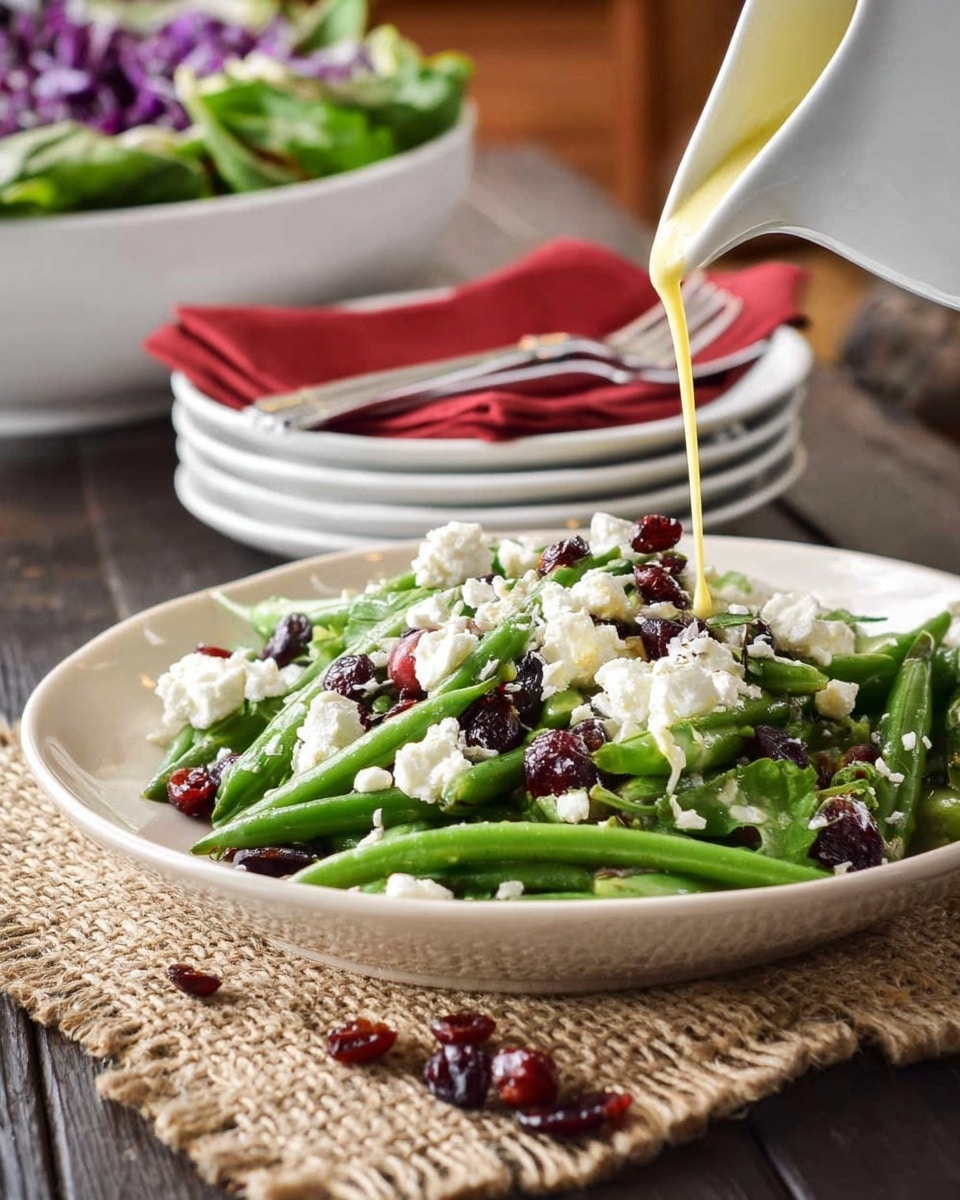 A white plate sits on a white marbled texture with a layer of bright green snap peas and green beans as the base, topped with crumbled white cheese scattered unevenly across the vegetables, and red dried cranberries sprinkled on top and around the plate. A white pitcher is pouring a light yellow dressing over the salad. In the background, a white bowl filled with green and purple salad is slightly out of focus, along with stacked white plates with a red napkin and silver cutlery resting on them. Photo taken with an iphone --ar 4:5 --v 7