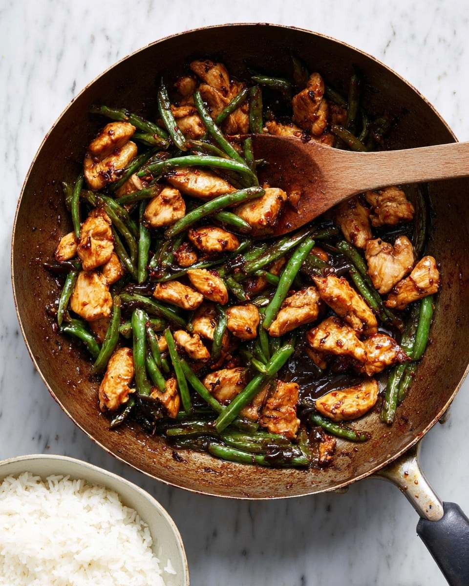 The image shows a frying pan filled with cooked chicken pieces and green beans stir-fried together. The chicken is golden brown with a slightly crispy texture, while the green beans are bright green and tender. The food looks coated with a dark, glossy sauce that adds shine and rich color. A wooden spoon rests inside the pan, partially covered with the sauce and vegetables. In the lower left corner, there is a white bowl filled with plain white rice. The whole scene is set on a white marbled surface. photo taken with an iphone --ar 4:5 --v 7