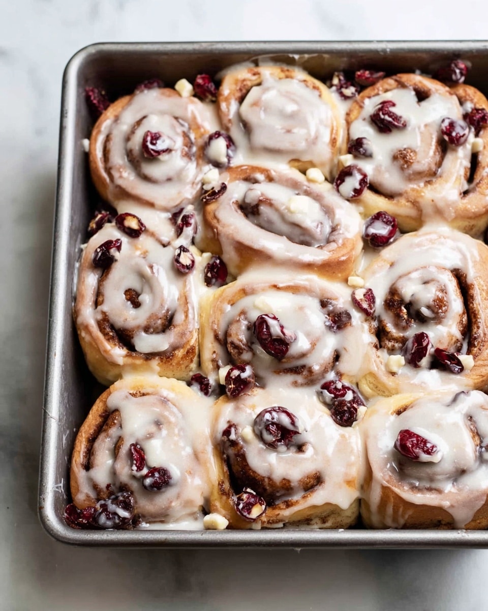 A metal rectangular baking pan filled with a dozen cinnamon rolls that have a light brown dough swirled with darker brown cinnamon filling, each roll sprinkled with dark red dried cranberries and dotted with small white chunks, all generously covered in creamy white icing that drapes over the rolls and pools slightly in spots, showing soft texture and a slightly glossy finish, placed on a white marbled surface photo taken with an iphone --ar 4:5 --v 7