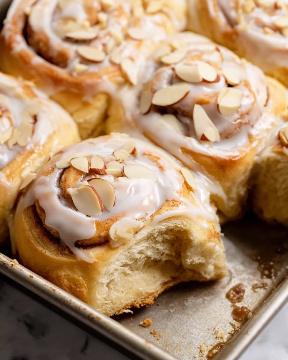 A close-up image showing four cinnamon rolls in a metal baking pan on a white marbled texture. Each cinnamon roll has a golden brown spiral dough base with a creamy white icing drizzled on top, creating smooth, shiny lines. Thin, pale beige almond slices are scattered over the icing, adding a slight texture contrast. The rolls have a soft, fluffy texture with visible layers of dough and cinnamon filling. One roll is slightly pulled away, revealing the soft, light brown inside and a moist, tender crumb. The pan’s surface shows some crumbs and syrup remnants. Photo taken with an iphone --ar 4:5 --v 7