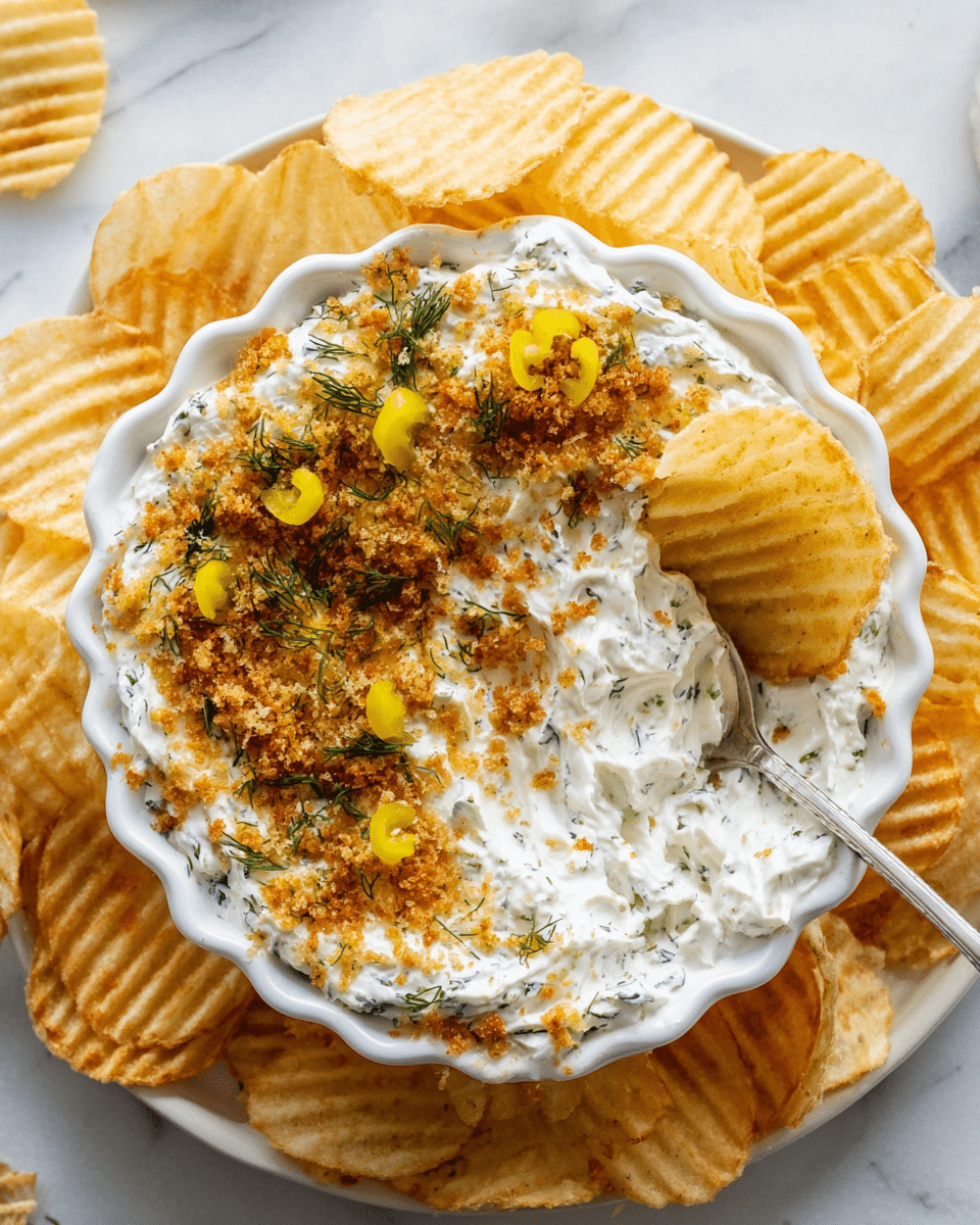 The image shows a white scalloped bowl filled with a creamy, thick white dip speckled with green herbs, topped with a layer of golden brown crispy crumbs and small bright yellow-green pickle pieces scattered on top. One ridged potato chip is partially dipped into the creamy dip on the right side of the bowl, and a silver spoon rests on the left side inside the bowl. The bowl sits on a round white plate, which is surrounded by ridged potato chips all around its edge. The background surface features a white marbled texture. photo taken with an iphone --ar 4:5 --v 7