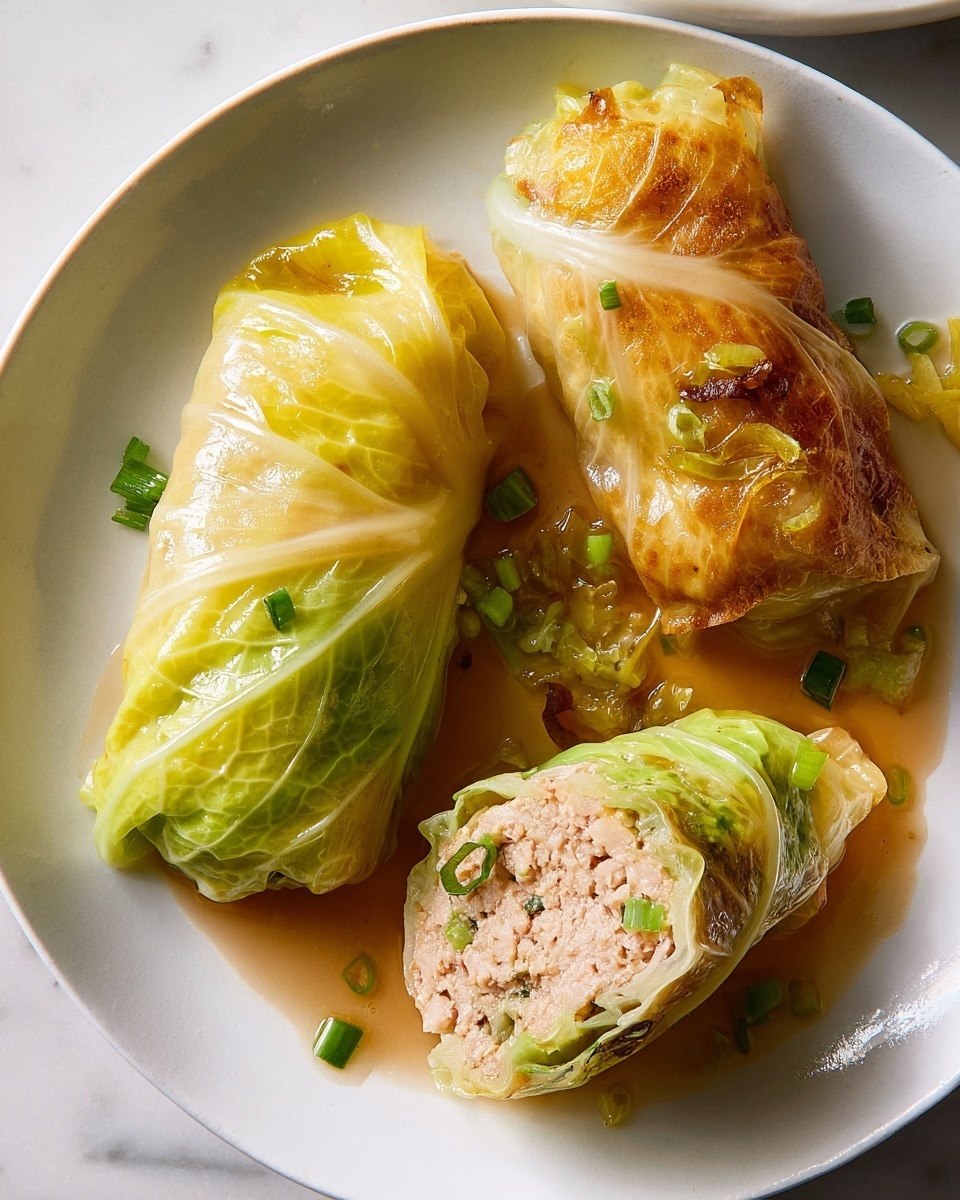 The image shows three cabbage rolls on a white plate with a white marbled background. Two whole cabbage rolls are placed side by side: one with a soft light green and yellow steamed cabbage leaf, the other with a darker golden-brown crispy cabbage leaf. A third cabbage roll is cut in half, showing the inner filling of light pink minced meat mixed with small green herbs. Scattered around the rolls are small pieces of chopped green onion and a shiny, light brown sauce giving a glossy texture to the dish. The lighting highlights the softness and crispy textures of the cabbage leaves, making the colors vibrant and appetizing. photo taken with an iphone --ar 4:5 --v 7
