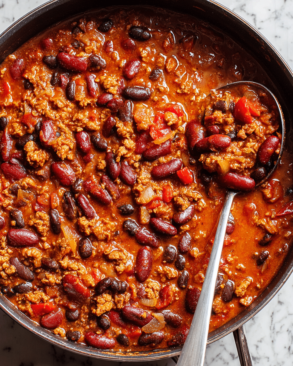 A close-up top view of a black pan filled with thick chili showing mixed layers of red kidney beans, dark black beans, and ground meat in a rich, orange-red sauce with visible chunks of onions and small bits of red peppers. A shiny metal ladle rests inside the pan, partially scooping some chili, displaying the chunky texture and glossy surface of the beans and meat, all set against a white marbled background. photo taken with an iphone --ar 4:5 --v 7