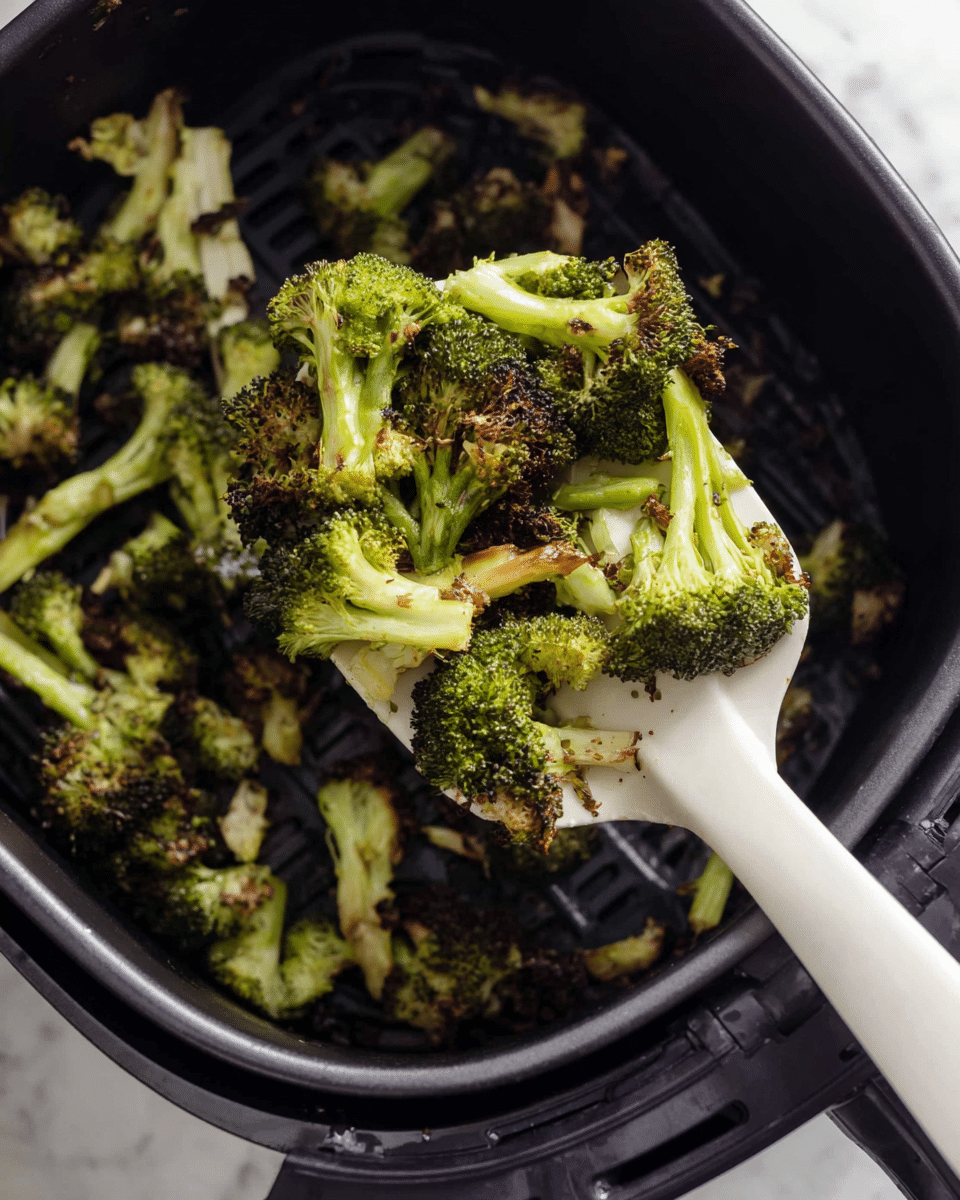 A close-up view of small, cooked broccoli pieces held on a white plastic spatula inside a black air fryer basket, showing a mix of bright green and browned, slightly crispy edges on the broccoli florets and stems, with a few leaves and small burnt bits scattered around the basket interior, all set on a white marbled surface. photo taken with an iphone --ar 4:5 --v 7