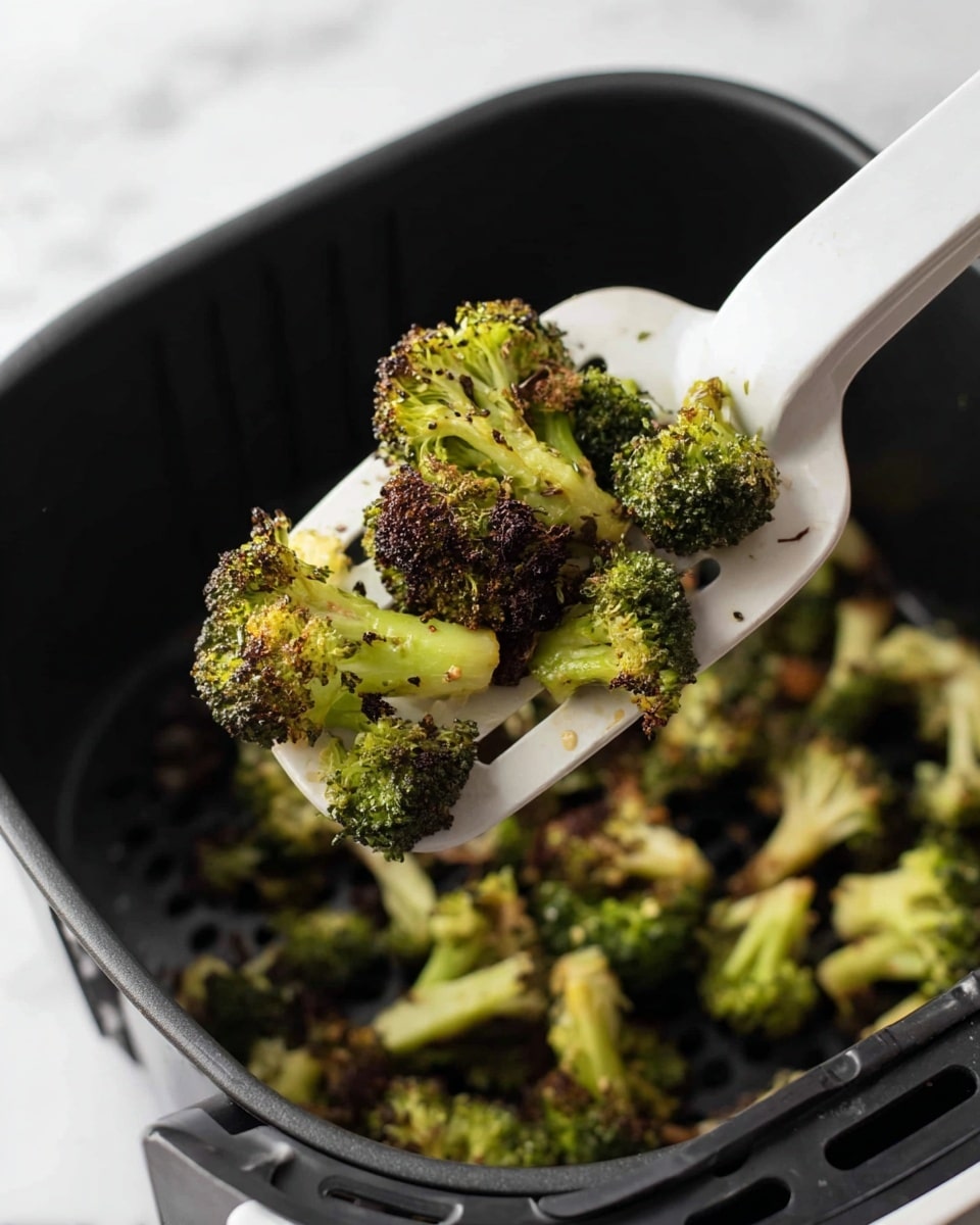 A close-up view of small pieces of roasted broccoli with a mix of green and dark brown colors, showing some parts charred and crispy, held on a white spatula above a black air fryer basket filled with more roasted broccoli. The broccoli pieces have a rough texture with distinct florets and some visible seasoning. The scene is set on a white marbled texture surface. photo taken with an iphone --ar 4:5 --v 7
