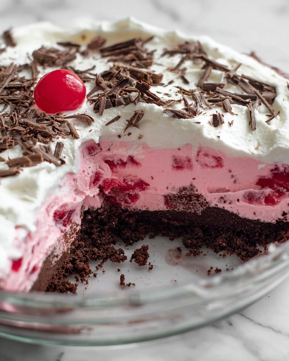 This image shows a layered dessert in a clear glass dish placed on a white marbled surface. The bottom layer is a thick, dark chocolate crumb crust with a rough texture. Above it is a bright pink creamy layer with bits of red fruit mixed in, looking soft and fluffy. The top layer is smooth white whipped cream, sprinkled generously with thin dark chocolate shavings, which add texture and color contrast. A single bright red cherry is placed on the left side of the dessert. A slice has been taken out showing the clear division between the layers. Photo taken with an iphone --ar 4:5 --v 7