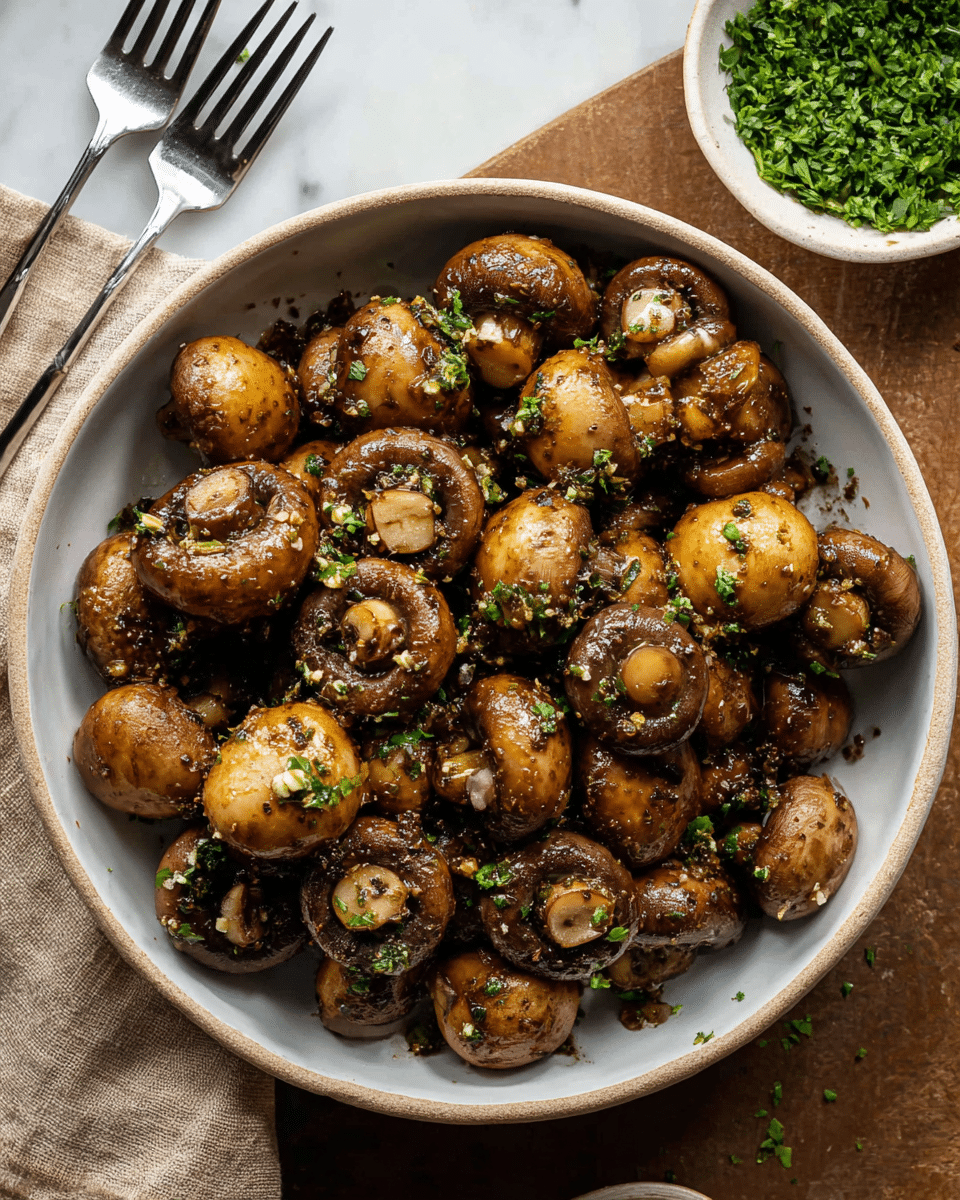 A deep white bowl is filled with cooked whole mushrooms that have a rich brown color, showing a shiny, oily texture with small bits of garlic and herbs coating them evenly. The mushrooms vary in size and appear tender with some showing their rounded tops and others showing the gills underneath. Small flecks of chopped green herbs are sprinkled throughout, adding a touch of green color. The bowl sits on a white marbled surface with a beige cloth and a fork nearby, and there is a small white bowl filled with more chopped green herbs in the top right corner. Photo taken with an iphone --ar 4:5 --v 7