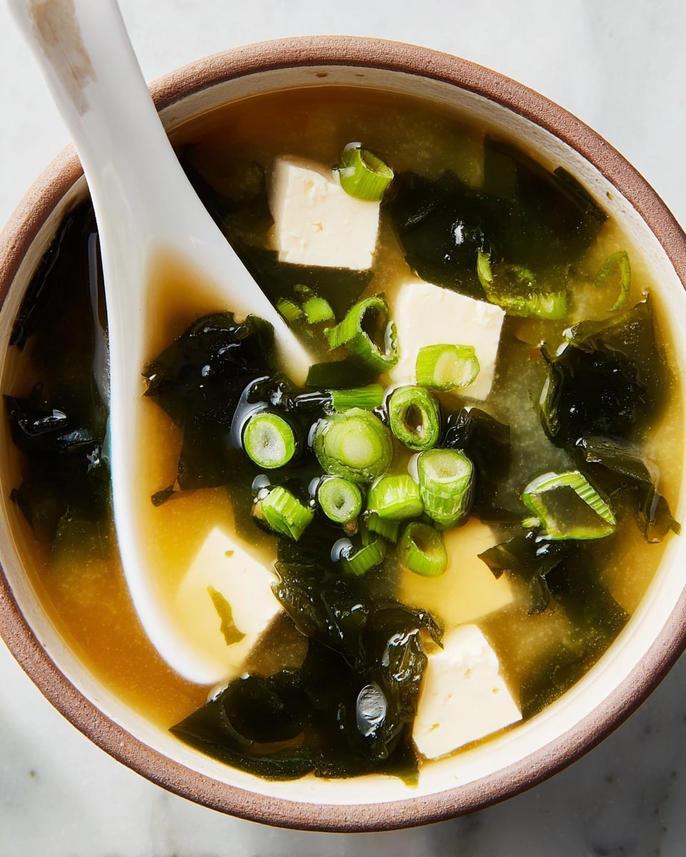 A close-up view of a bowl of clear light brown miso soup is shown, filled with dark green seaweed pieces floating on the surface. Inside the bowl, there are several small white tofu cubes with smooth, soft textures scattered among the seaweed. Bright green sliced scallions are sprinkled on top, adding a fresh pop of color against the broth. A white ceramic spoon with a slightly stained tip rests inside the bowl, partially submerged in the soup. The bowl is placed on a white marbled surface. photo taken with an iphone --ar 4:5 --v 7
