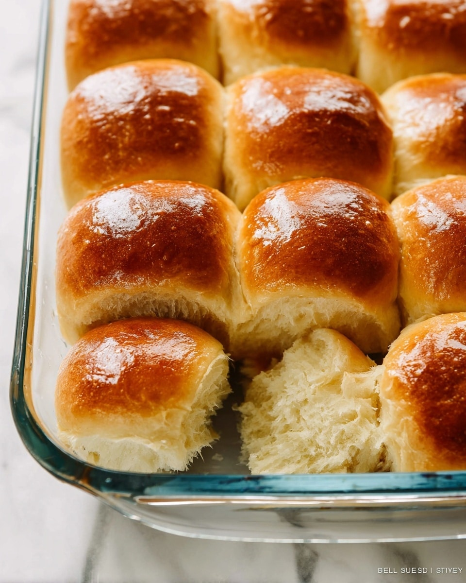 A close-up view of a glass baking dish filled with nine soft, golden-brown dinner rolls arranged in three rows of three. Each roll has a shiny, slightly textured surface from being brushed with a white glaze using a paintbrush held by a woman's hand in the top right corner. The rolls touch each other, creating a fluffy yet compact look. The background is a white marbled surface, enhancing the warm tones of the buns. Photo taken with an iphone --ar 4:5 --v 7