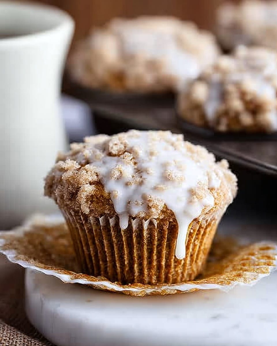 A single crumb-topped muffin with a light brown color sits in a white paper liner, partially peeled open at the base, covered by a smooth white glaze that drips unevenly down the sides. Behind it, a dark metal tray holds several more muffins with the same crumbly texture, all resting on a white marbled surface. To the left, part of a white cup is visible, adding contrast to the warm-toned muffin. Photo taken with an iphone --ar 4:5 --v 7