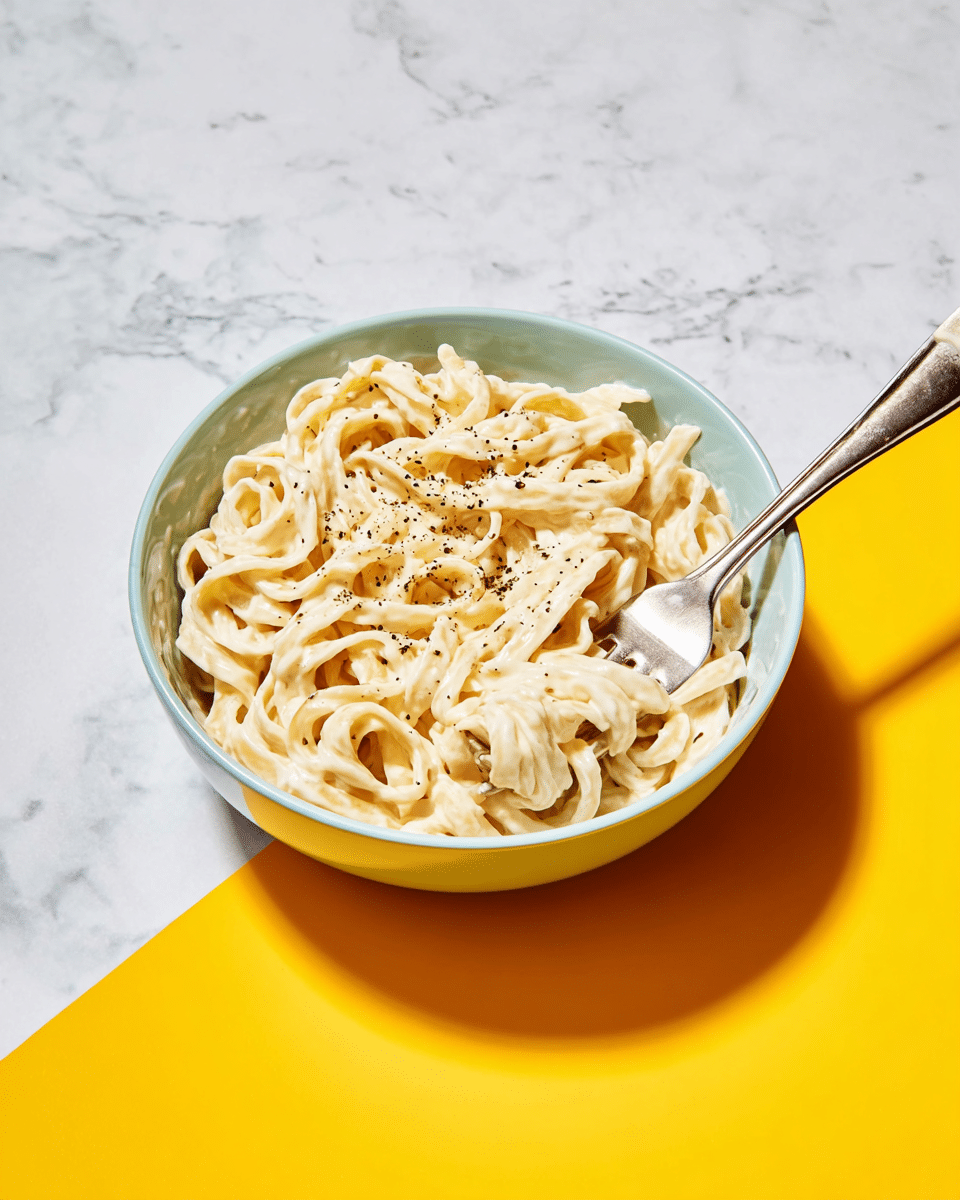 A white bowl filled with creamy fettuccine pasta, coated evenly in a smooth white sauce, with visible small black pepper flakes sprinkled on top. The pasta noodles are thick and twisted, filling the bowl fully, with a silver fork inserted on the right side, slightly lifting some noodles. The bowl is placed on a surface that is half white marbled texture and half bright yellow. The lighting is bright and highlights the creamy texture of the sauce and the pepper details. photo taken with an iphone --ar 4:5 --v 7