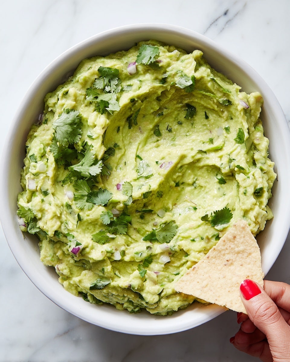 A white bowl filled with two layers of creamy guacamole, the base layer smooth and light green with some darker green cilantro leaves mixed throughout, and the top layer slightly chunkier with small bits of purple onion and green herbs visible. Fresh cilantro leaves are scattered on top as garnish. On the right side of the bowl, a woman's hand with red nail polish holds a single triangular light beige tortilla chip, partially dipped into the guacamole. The bowl sits on a white marbled surface. photo taken with an iphone --ar 4:5 --v 7