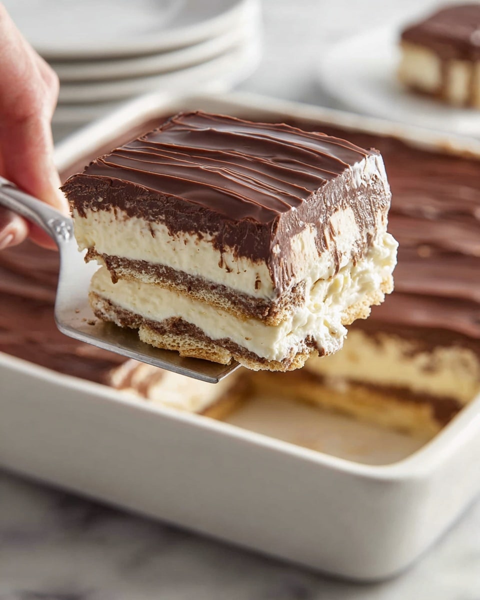 A square slice of layered dessert is lifted by a spatula above a white baking dish on a white marbled surface. The dessert has three main layers: a thick, glossy dark brown chocolate top layer with a smooth, slightly wavy texture; beneath that, two thick creamy beige layers that look soft and smooth, separated by thin layers of light brown crunchy cookie or wafer. The layers of cookie or wafer are visible inside and at the base, adding a firm texture. A woman's hand holds the spatula, showing the dessert's size and thickness. In the background, part of the uncut dessert with the same layered texture is visible. Photo taken with an iphone --ar 4:5 --v 7