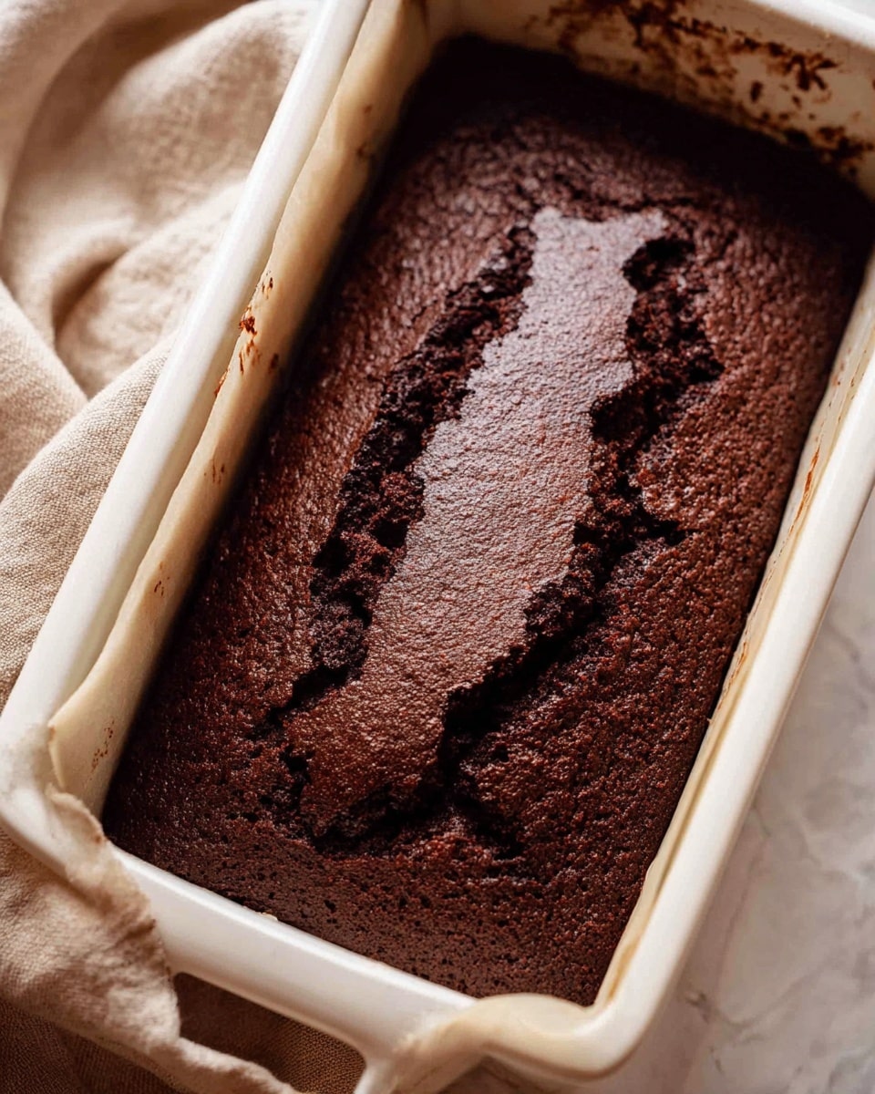 A close-up image of a freshly baked dark chocolate cake with a rough, cracked top layer showing a dense texture, all inside a white loaf pan lined with parchment paper. The sides of the pan have faint stains, and the whole setup is placed on a white marbled textured surface with a soft beige cloth peeking from the top left corner. The cake’s surface is matte with uneven cracks revealing a slightly moist inside. Photo taken with an iphone --ar 4:5 --v 7