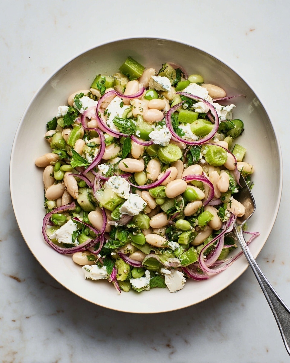 A white bowl filled with a bean salad showing one main layer of white beans mixed with bright green chopped celery and fresh green herbs, scattered pieces of soft white cheese, and thin slices of purple-red onion curls mixed throughout. A silver spoon rests inside the bowl, partially buried in the salad. The bowl is set on a white marbled surface. photo taken with an iphone --ar 4:5 --v 7