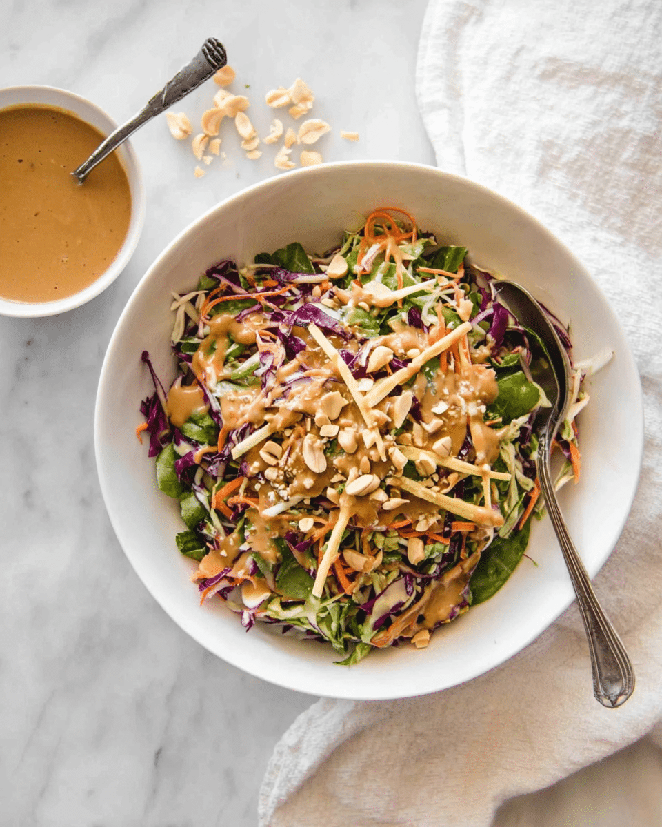 A white bowl filled with a colorful salad made of thin purple cabbage strands, green leafy vegetables, and shredded orange carrots, topped with crunchy light brown chopped nuts and thin crunchy sticks of pale yellow color. The salad is drizzled with a creamy brown dressing that is also seen in a small white bowl on the side, with a spoon resting in it. A silver spoon is placed inside the bowl, partially covered by the salad. The bowl is placed on a white marbled surface with a white cloth nearby. Photo taken with an iphone --ar 4:5 --v 7