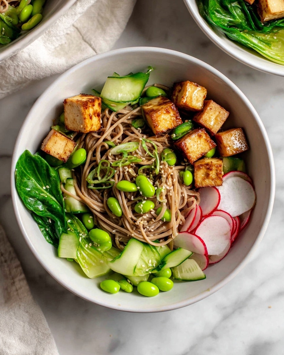 The image shows a white bowl filled with a base layer of light brown soba noodles in the center. On top of the noodles are green edamame beans scattered evenly. Around the middle and towards the edges are golden-brown crispy tofu cubes. The bowl’s edge is lined with dark green cooked baby bok choy leaves, fresh light green cucumber slices, and thin red and white radish slices. The bowl sits on a white marbled surface with part of another white bowl visible in the background. Photo taken with an iphone --ar 4:5 --v 7
