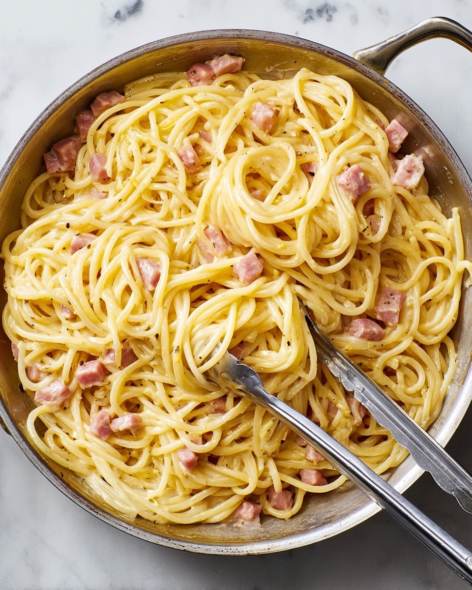 A round pan filled with creamy yellow spaghetti pasta mixed with small pieces of light pink ham, showing a smooth and glossy texture. The spaghetti is piled loosely with some strands curling over each other, and the ham pieces are spread evenly throughout. Two silver tongs rest inside the pan, partially buried in the pasta. The pan sits on a white marbled surface with a clean and simple look. photo taken with an iphone --ar 4:5 --v 7