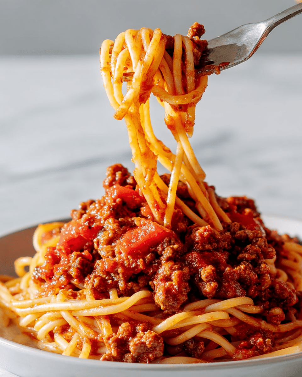 A close-up of a white plate piled with spaghetti mixed with a chunky red meat sauce, showing layers of thick noodles intertwined with pieces of browned ground meat and diced tomatoes, with a fork lifting a tangle of spaghetti above the plate, the sauce coating the noodles richly, all set against a white marbled background. photo taken with an iphone --ar 4:5 --v 7