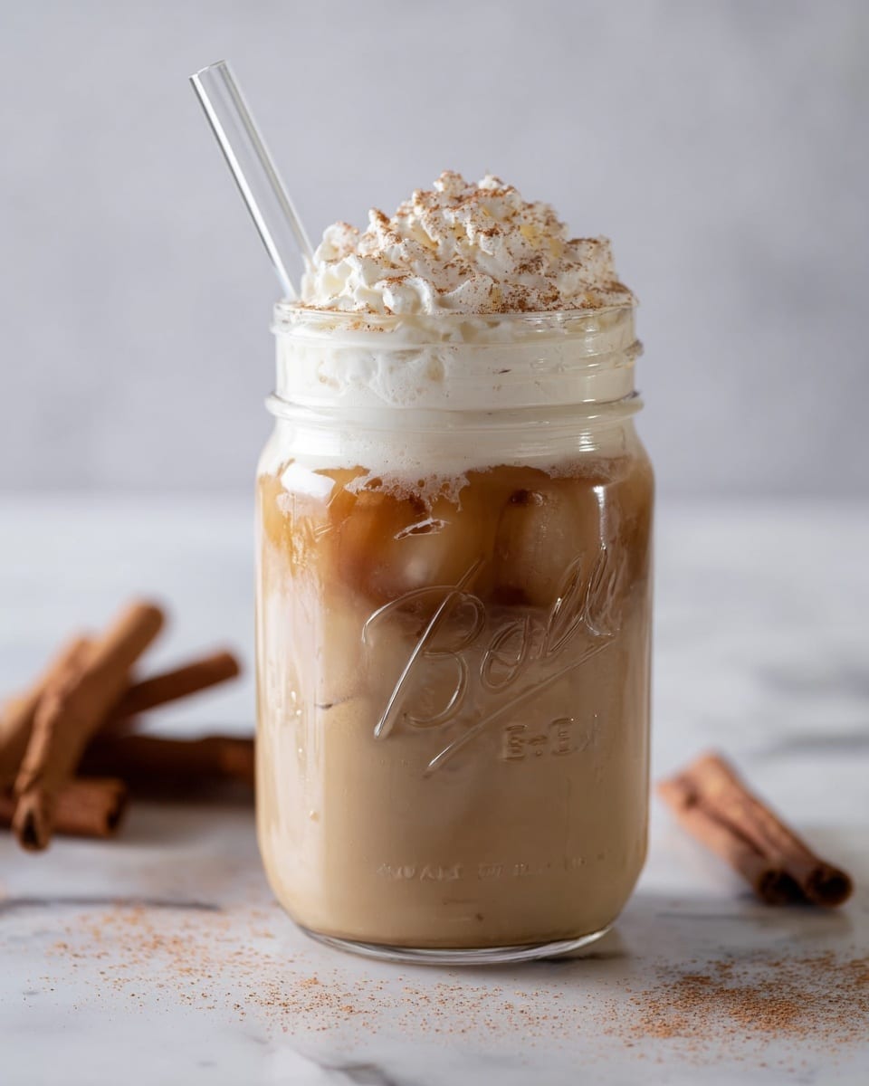 A clear glass jar filled with three visible layers: the bottom layer is a light brown creamy liquid, the middle layer shows slightly darker coffee mixed with ice cubes, and the top layer is white whipped cream sprinkled with light brown cinnamon powder; a clear straw is inserted into the jar from the top left side, all placed on a white marbled surface with some cinnamon sticks nearby. photo taken with an iphone --ar 4:5 --v 7