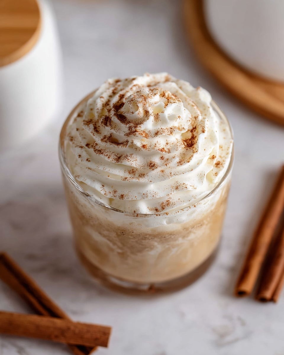 A glass cup filled with a creamy light brown drink at the base, topped with a thick layer of white whipped cream swirled on top, sprinkled with fine brown cinnamon powder evenly spread over the whipped cream. The cup is placed on a white marbled surface with some cinnamon sticks blurred in the background near the top left and bottom right edges. A white container with a wooden lid is also blurred at the bottom center. Photo taken with an iphone --ar 4:5 --v 7