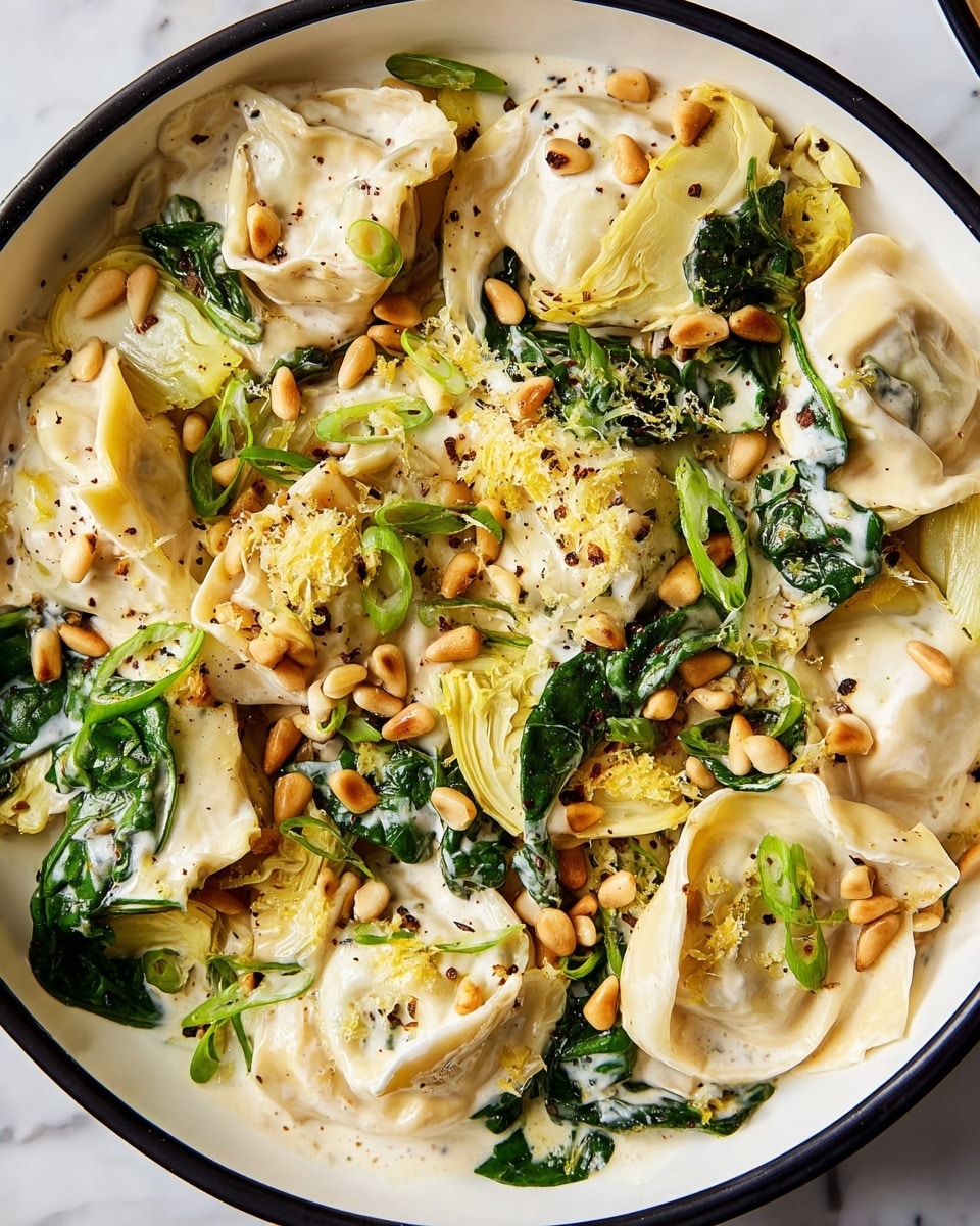 This dish features a single layer of tortellini pasta coated in a creamy white sauce, scattered with small toasted pine nuts that are golden brown. Mixed in are dark green spinach leaves and pale yellow artichoke pieces, creating a mix of colors and textures. The pasta is sprinkled with thin slices of bright green spring onions and black pepper, with a few tiny yellow lemon zest bits adding brightness. A silver fork with a wooden handle rests on the edge of a white plate. The plate sits on a white marbled surface. Photo taken with an iphone --ar 4:5 --v 7
