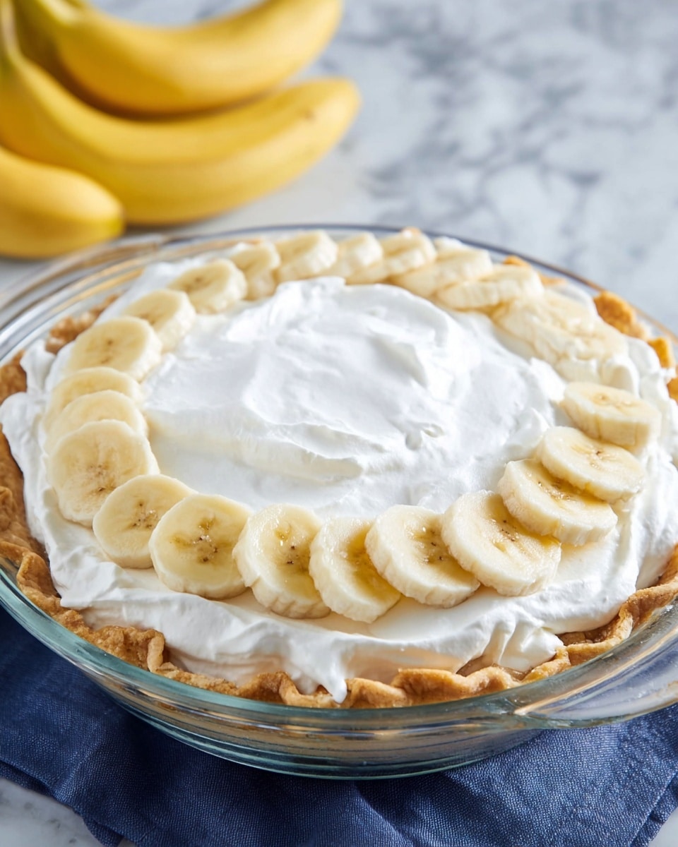 A clear glass pie dish holds a pie with three visible layers: a golden brown flaky crust with folded edges on the bottom, a thick layer of smooth white whipped cream covering most of the surface, and a ring of sliced yellow bananas with a slightly light brown center placed evenly around the pie's edge on top of the whipped cream. The pie dish sits on a white marbled surface with a blue cloth underneath. Two whole bananas lie in the background. Photo taken with an iphone --ar 4:5 --v 7