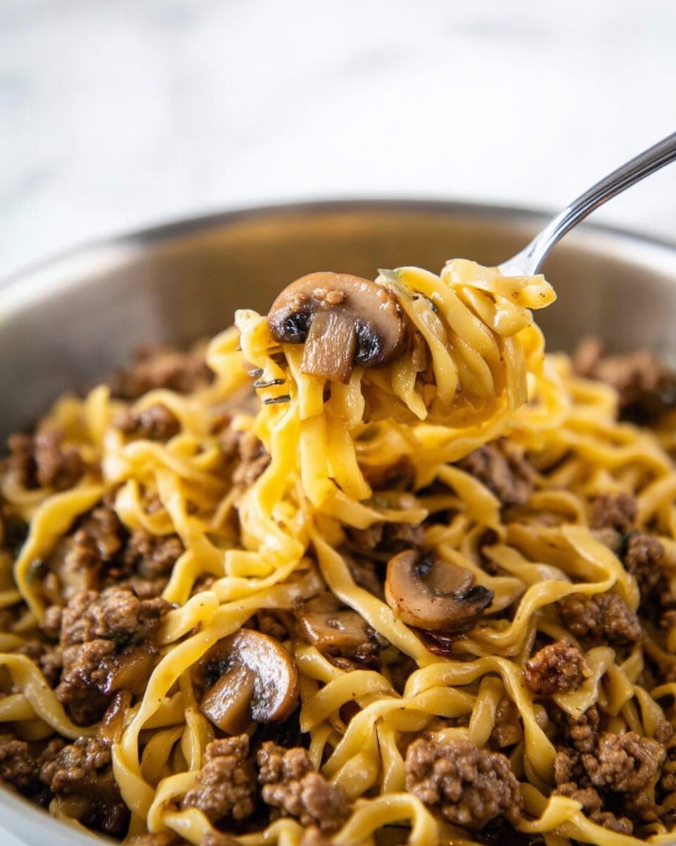Close-up of a white bowl filled with yellow egg noodles mixed with small pieces of brown cooked ground meat and sliced brown mushrooms. A silver fork holds a bite of the noodles, meat, and mushroom lifted above the bowl. The background is a white marbled texture. The noodles are soft and slightly shiny, the meat has a crumbly texture, and the mushrooms appear tender and moist. Photo taken with an iphone --ar 4:5 --v 7