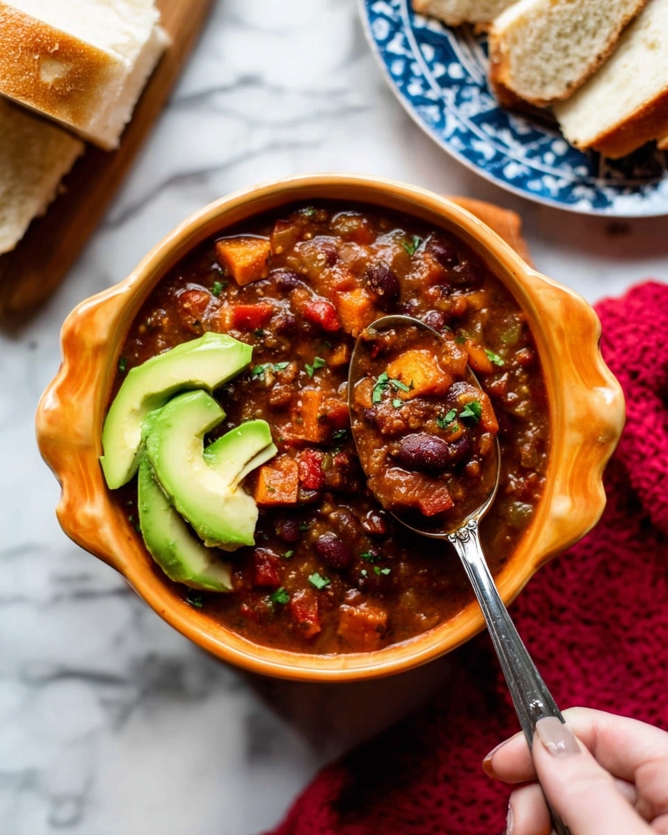 A bowl of thick chili with dark red beans, browned chunks of tofu, and diced orange and red vegetables in a rich brown sauce fills a scalloped orange bowl. On top, two slices of fresh green avocado rest as garnish. A woman's hand holds a silver spoon scooping into the chili, showing the thick texture with visible vegetable pieces and herbs. The bowl sits on a white marbled surface with a blue patterned plate holding sliced white bread in the background and a red cloth nearby, adding warm color contrast. Photo taken with an iphone --ar 4:5 --v 7