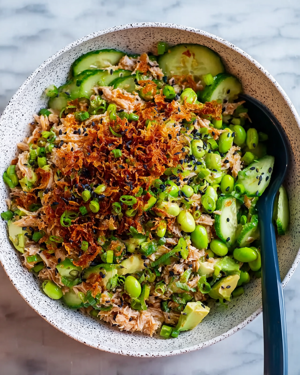 A white bowl with black speckles holds a colorful mixed dish with several layers. The bottom layer is light brown shredded cooked tuna spread evenly. On top, there are many slices of green cucumber mixed with bright green edamame beans and light green avocado pieces. Scattered throughout are finely chopped green onions. Over the entire dish, there is a drizzle of reddish-brown crispy fried onions and small black sesame seeds sprinkled. A dark blue spoon is placed inside the bowl on the right side. The bowl rests on a white marbled surface. Photo taken with an iphone --ar 4:5 --v 7