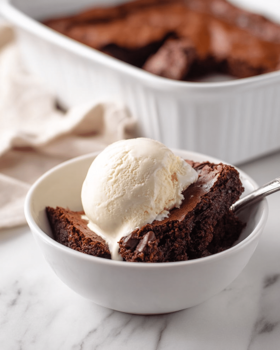 A white bowl holds a rich, dark brown chocolate brownie piece that looks soft and slightly cracked on top, with a smooth, round scoop of creamy vanilla ice cream placed on it. The background shows a white baking dish filled with more brownies, slightly out of focus. The scene is set on a white marbled surface, adding a clean and fresh look. photo taken with an iphone --ar 4:5 --v 7