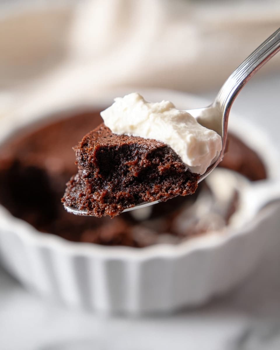 A close-up view of a silver spoon holding a scoop of dark brown, moist chocolate cake with a light layer of creamy white frosting underneath; the background shows a white fluted bowl filled with more of the chocolate cake, set on a white marbled surface with soft natural light. photo taken with an iphone --ar 4:5 --v 7