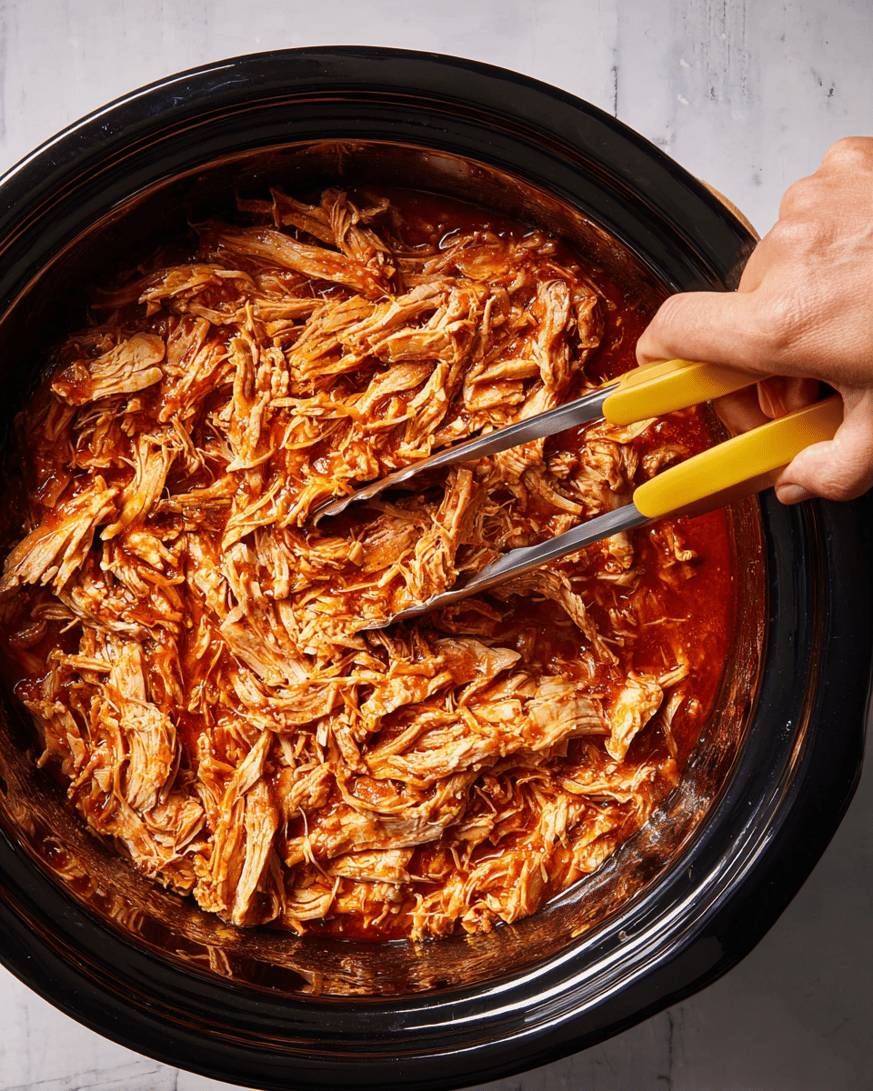 A close-up view of shredded chicken soaked in a thick, reddish-brown sauce, filling a large black slow cooker. The chicken is tender and stringy, layered evenly to cover the whole area inside the cooker. On the right side, a woman's hand is holding yellow-handled tongs, ready to pick up the chicken. The background has a white marbled texture. Photo taken with an iphone --ar 4:5 --v 7