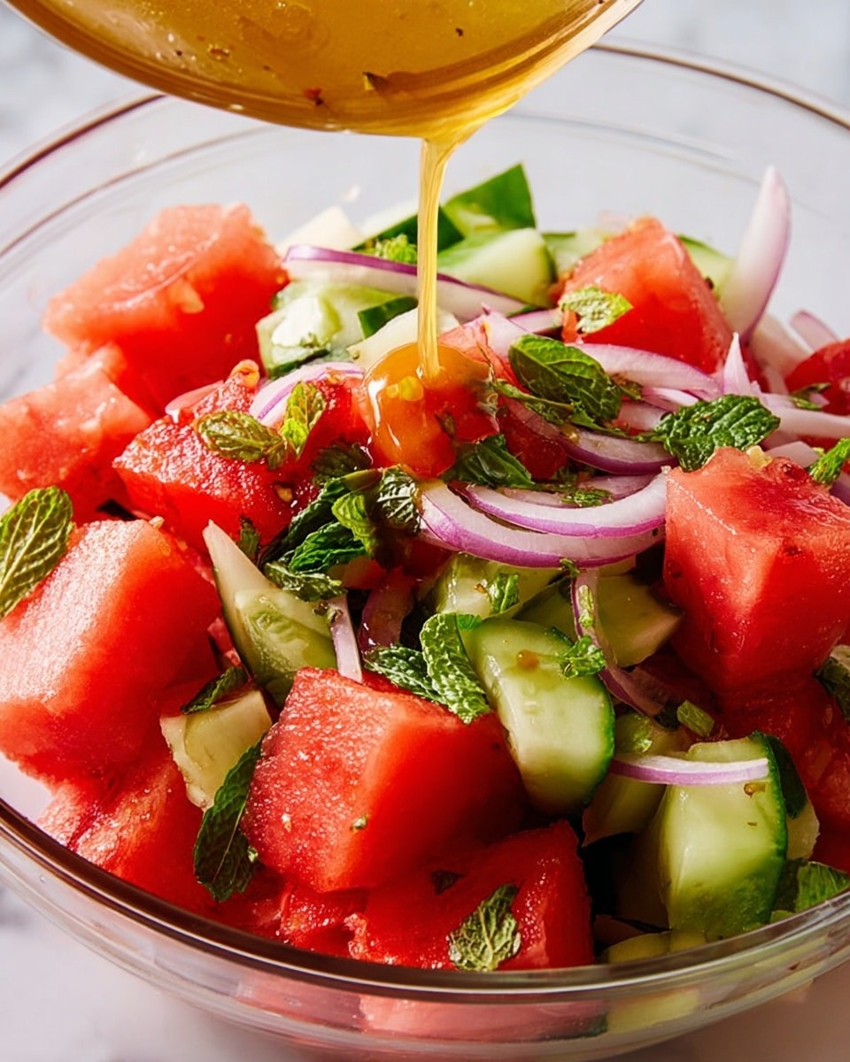 A close-up view of a clear glass bowl filled with three main layers of fresh salad ingredients: bright red watermelon cubes, light green cucumber pieces, and thin slices of purple onion, all mixed with small green mint leaves scattered throughout. A golden yellow dressing is being poured over the salad from above, adding a shiny texture to the top. The bowl sits on a white marbled texture surface. photo taken with an iphone --ar 4:5 --v 7