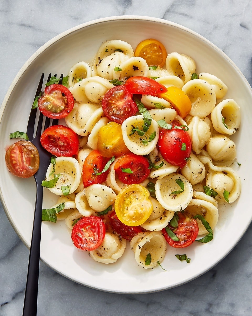 The image shows a white round plate filled with two layers of food: the bottom layer contains small, round, pale yellow orecchiette pasta, and the top layer consists of halved red and yellow cherry tomatoes along with small green basil leaves scattered throughout. The pasta and tomatoes have a light coating of oil and black pepper sprinkled on top, giving a slight shine and texture. A black fork rests on the left side of the plate, holding a couple of pasta pieces and tomato halves. The plate is placed on a white marbled surface. photo taken with an iphone --ar 4:5 --v 7