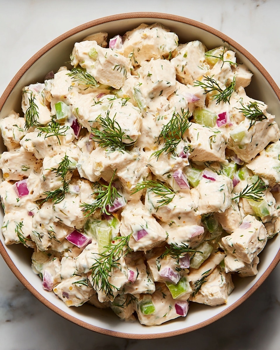 A close-up view of a creamy chicken salad served in a white bowl with a tan rim, placed on a white marbled surface. The salad has three main parts: chunks of white cooked chicken coated in a smooth, pale beige dressing, small pieces of green celery and bright purple-red onion mixed throughout, and garnished with delicate sprigs of fresh green dill scattered all over. The textures show both the soft creaminess of the dressing and the crunchy bits of vegetable. Photo taken with an iphone --ar 4:5 --v 7