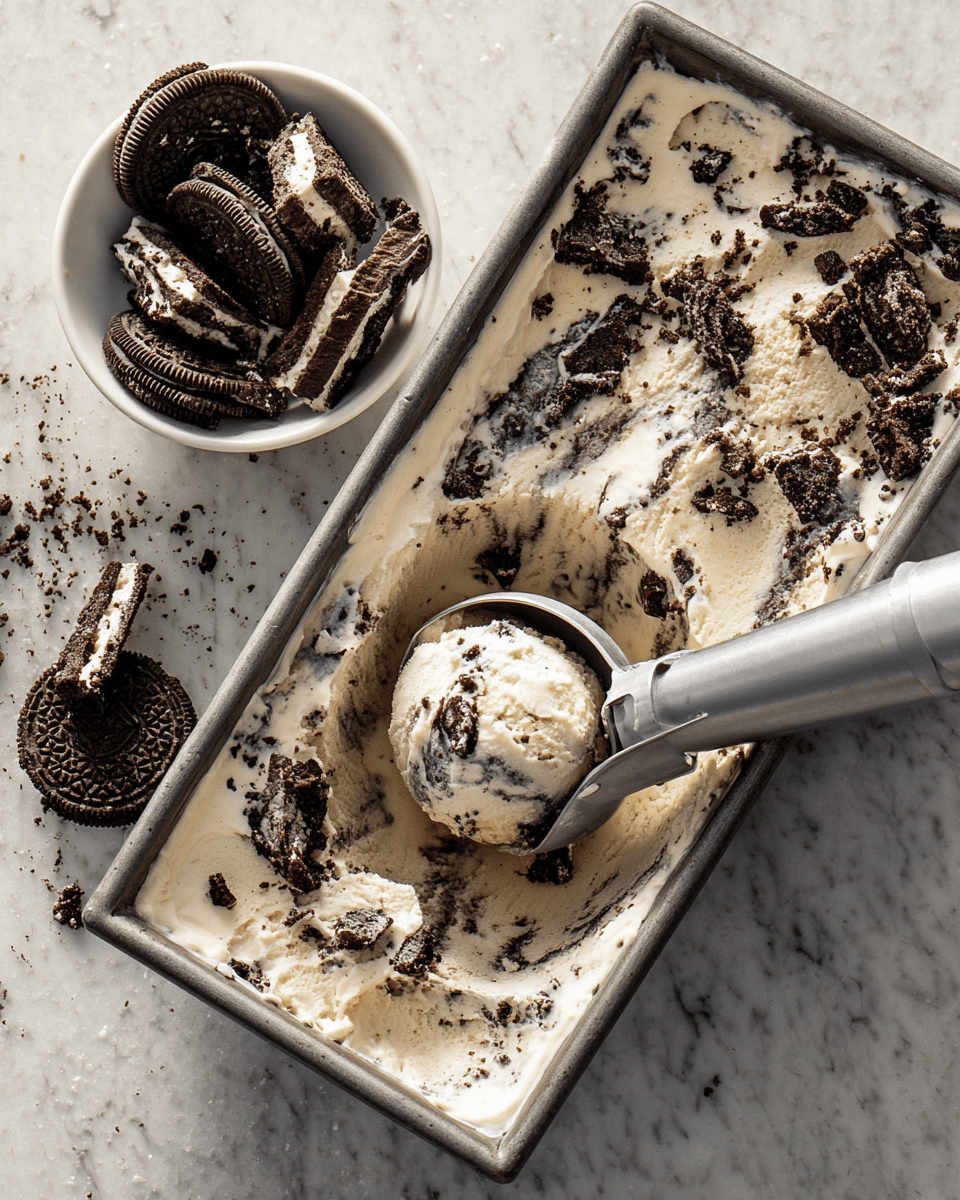A metal rectangular container holds creamy vanilla ice cream mixed with many chunks of dark chocolate cookies throughout the single thick layer. On the side, a white bowl contains more broken dark chocolate cookies with white filling, sitting on a white marbled surface. A silver ice cream scooper is digging into the ice cream, lifting a rounded scoop filled with cookie pieces. The ice cream looks soft and slightly melting, and the cookie pieces add a rough, crumbly texture scattered on top. photo taken with an iphone --ar 4:5 --v 7