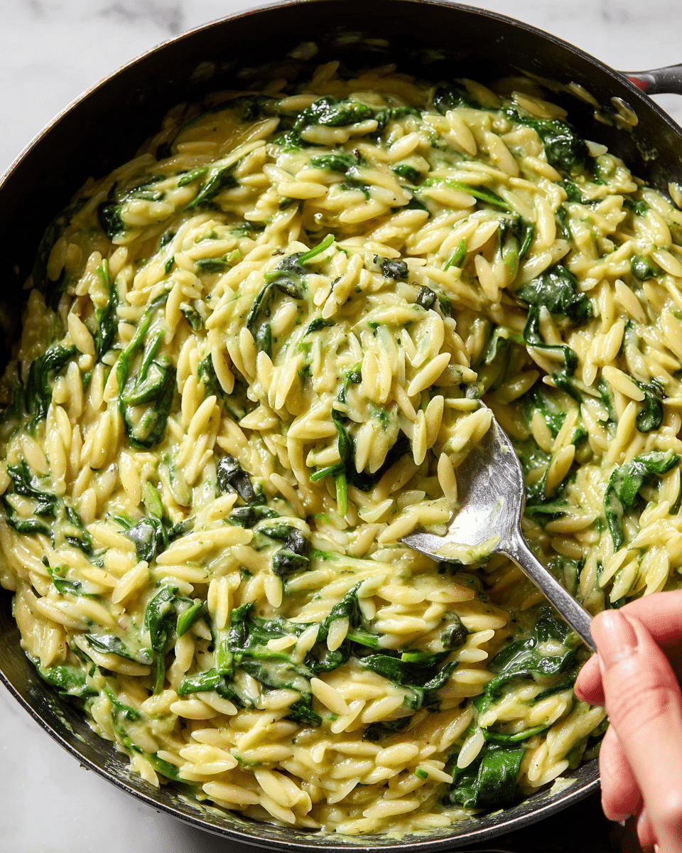 A close-up view of a creamy yellow-green orzo pasta dish mixed with bright green spinach leaves in a black pan, showing thick sauce clinging to the pasta and the wilted spinach scattered evenly throughout. A woman's hand is holding a shiny silver spoon scooping up a portion of the orzo from the right side, revealing a textured, rich, and smooth consistency. The background is a white marbled surface, giving a clean and simple look. photo taken with an iphone --ar 4:5 --v 7