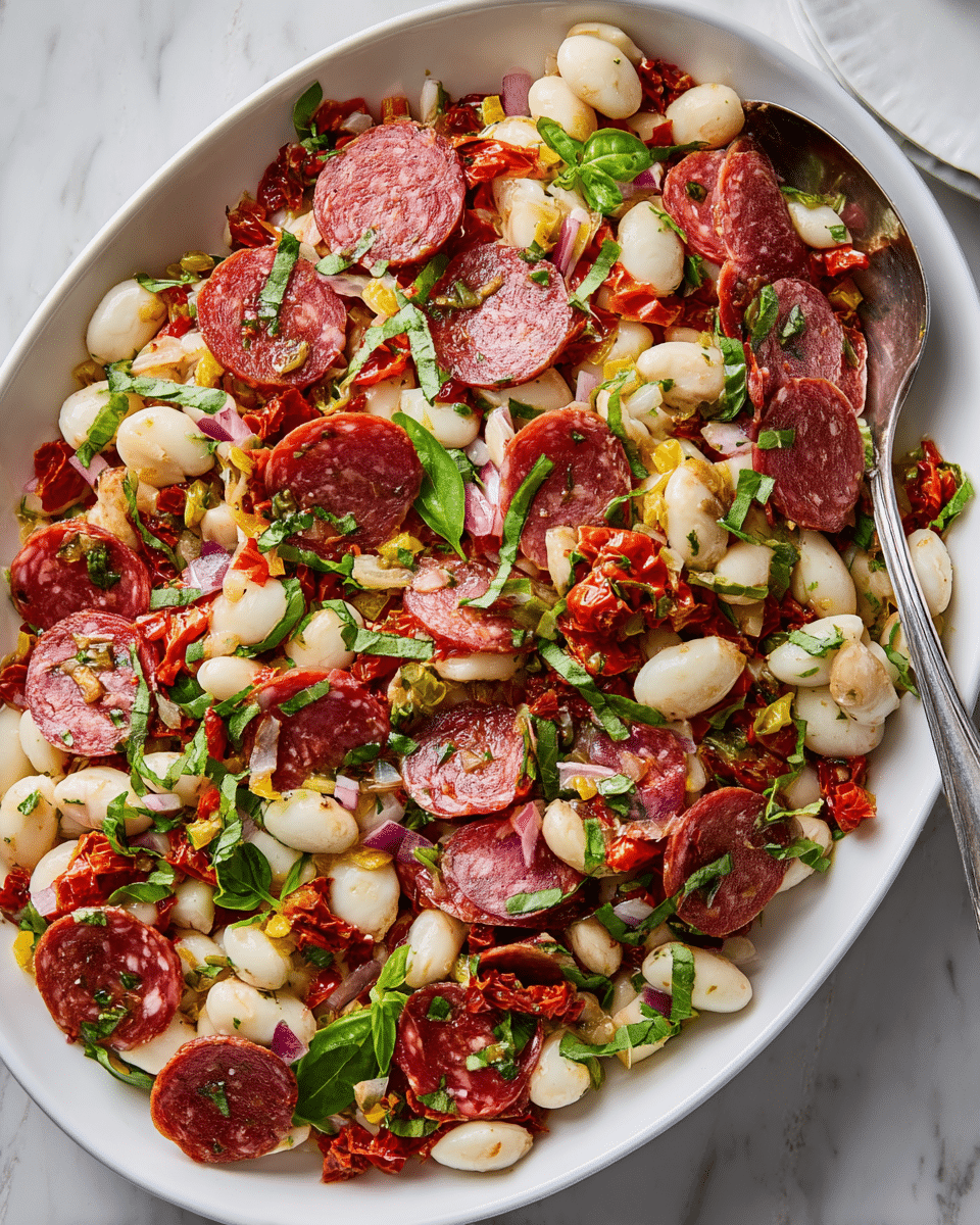 A white oval dish filled with a mixed salad showing layers of creamy white beans and small white mozzarella balls as the base, topped with thin reddish-pink slices of salami scattered evenly. Mixed throughout are small pieces of diced red onions, tiny bright yellow pepper bits, and finely chopped green basil leaves sprinkled on top. The salad also contains diced sun-dried tomatoes adding red specks. A silver spoon lies partially inside the dish against a white marbled surface background. photo taken with an iphone --ar 4:5 --v 7